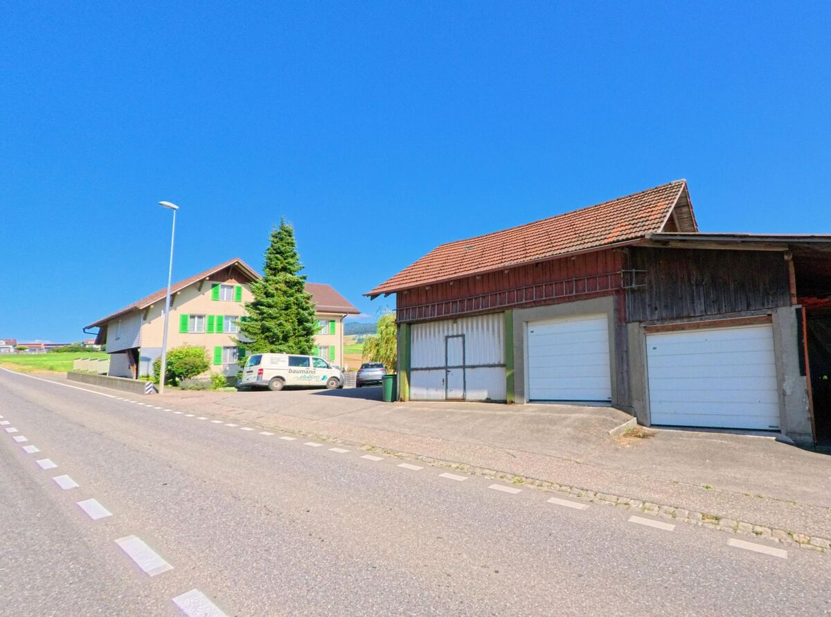 In Freimettigen (3510, Kanton Bern) verlaufen Wohnhaus und Scheune mit Garagen entlang einer leicht ansteigenden Hauptstrasse, gesaeumt von einem Nadelbaum und abgestellten Fahrzeugen.