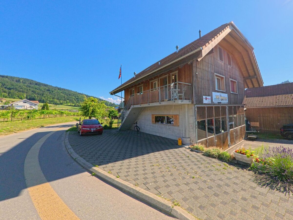 In Freimettigen (3510, Kanton Bern) befindet sich ein modernes Holzhaus mit Schweizer Fahne und grossem gepflastertem Vorplatz an einer Kurve mit Blick auf Reben und Huegel.