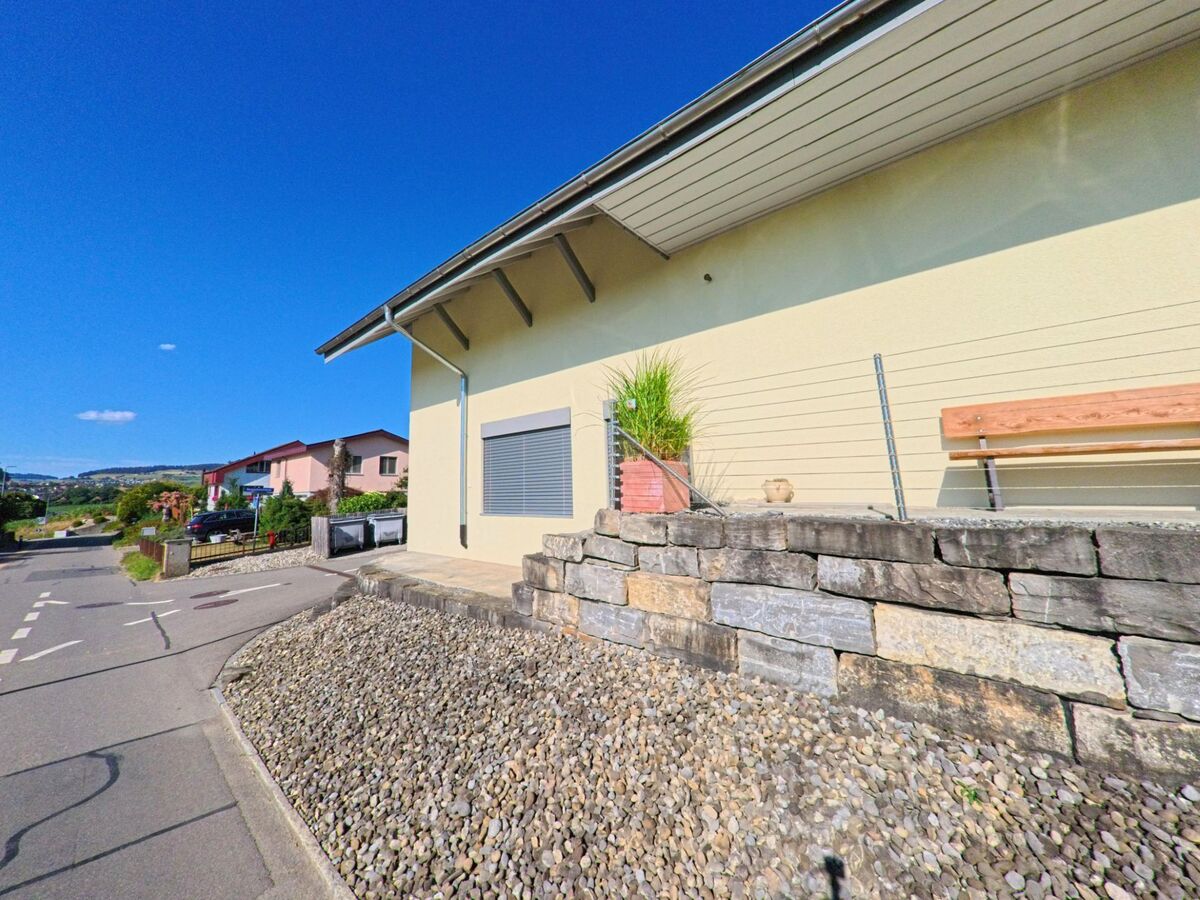 In Freimettigen (3510, Kanton Bern) fuehrt eine Strasse an einem modernen Gebaeude mit gelber Fassade vorbei, dessen vorgelagerte Steinmauer eine Sitzbank und Pflanzkübel traegt.