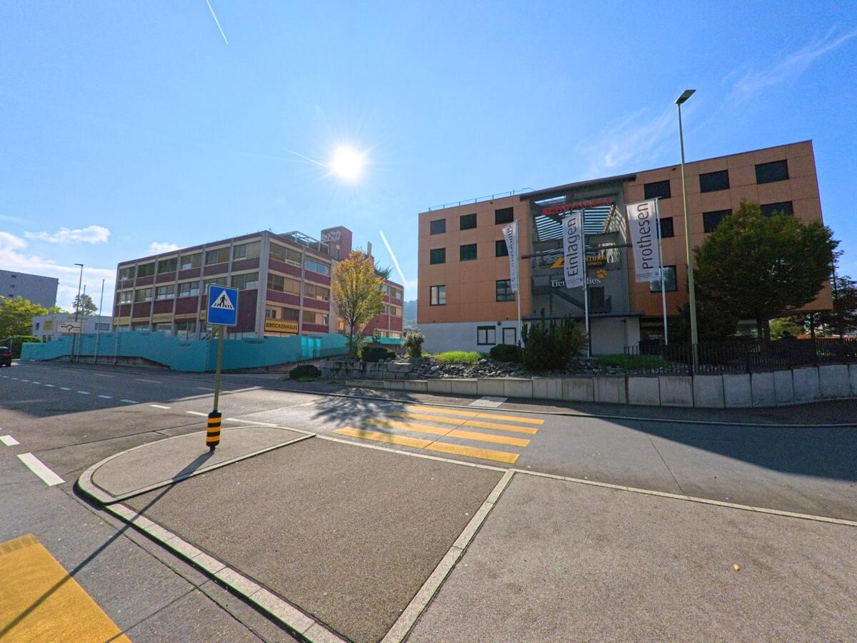 Gegenueber einem Fussgaengerstreifen in Frenkendorf (4402, Kanton Basel-Landschaft) stehen zwei gewerbliche Gebaeude, eines mit farbiger Fassade und das andere mit grossen Werbebannern.