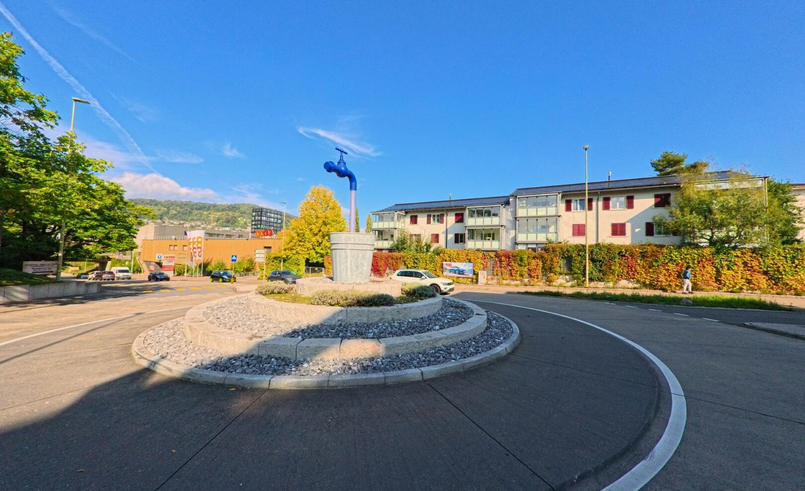 Ein Kreisverkehr in Frenkendorf (4402, Kanton Basel-Landschaft) zeigt eine markante blaue Armatur als zentrales Kunstobjekt vor Wohnhaeusern und herbstlicher Begruenung.
