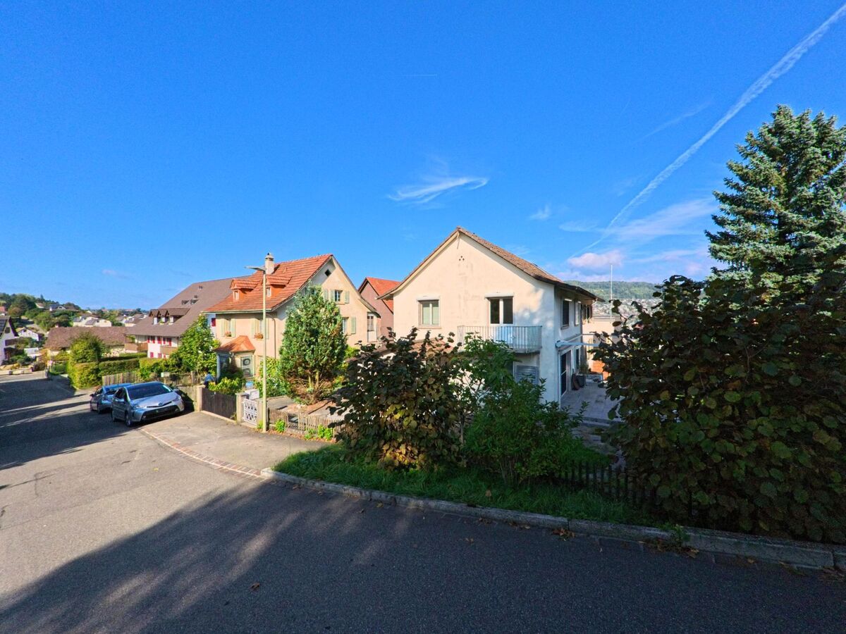 In einem Wohnquartier von Frenkendorf (4402, Kanton Basel-Landschaft) reihen sich Einfamilienhaeuser mit Gaerten an einer leicht abfallenden Strasse unter blauem Himmel.