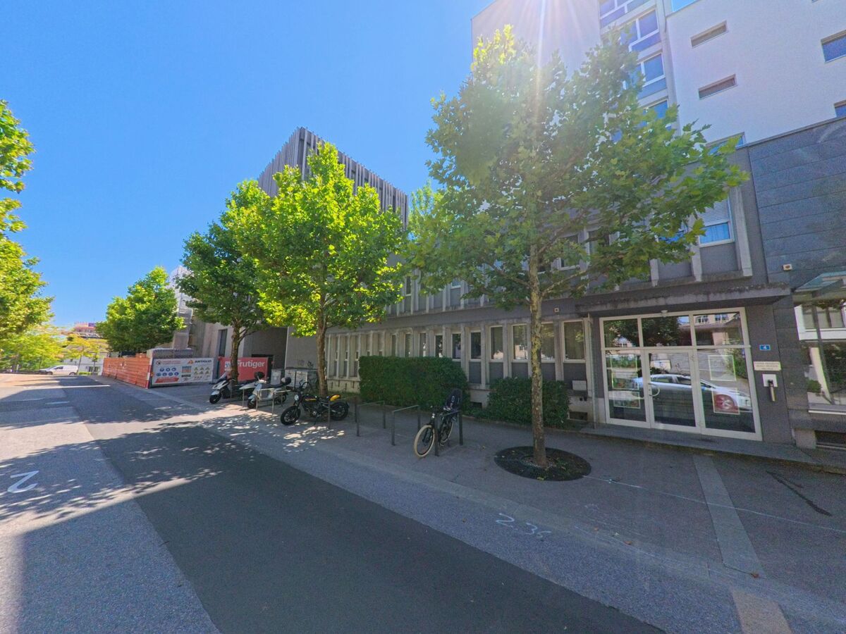 Vor einem modernen Gebaeude in Fribourg (1700, Kanton Freiburg) stehen mehrere Fahrraeder und Motorraeder entlang einer von Baeumen gesaeumten Strasse.