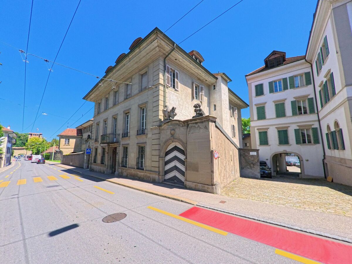 Ein historisches Stadthaus mit markantem Tor in Fribourg (1700, Kanton Fribourg) steht an einer Strasse mit Zebrastreifen und angrenzenden Wohnhaeusern.