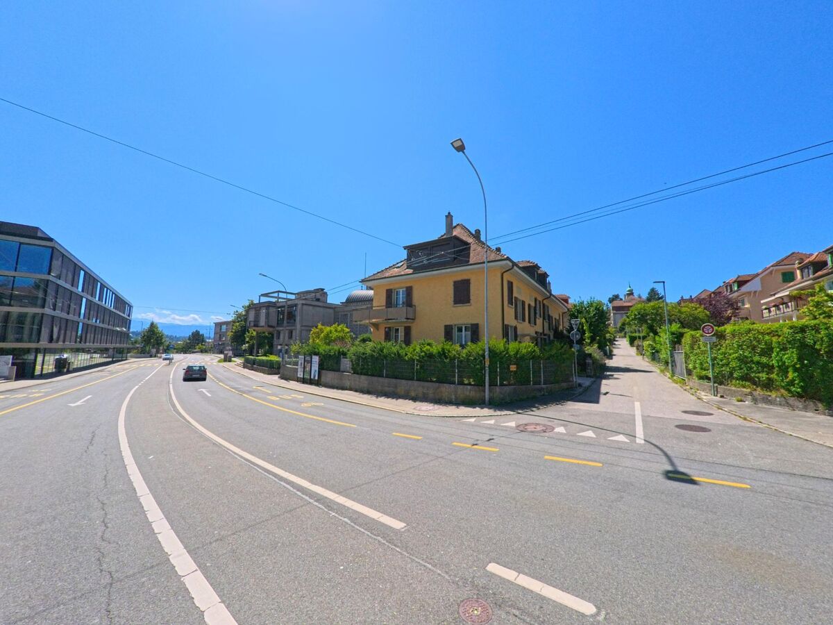Eine breite Strasse in Fribourg (1700, Kanton Fribourg) verläuft zwischen modernen Gebaeuden und Wohnhaeusern, im Hintergrund sind die Alpen sichtbar.