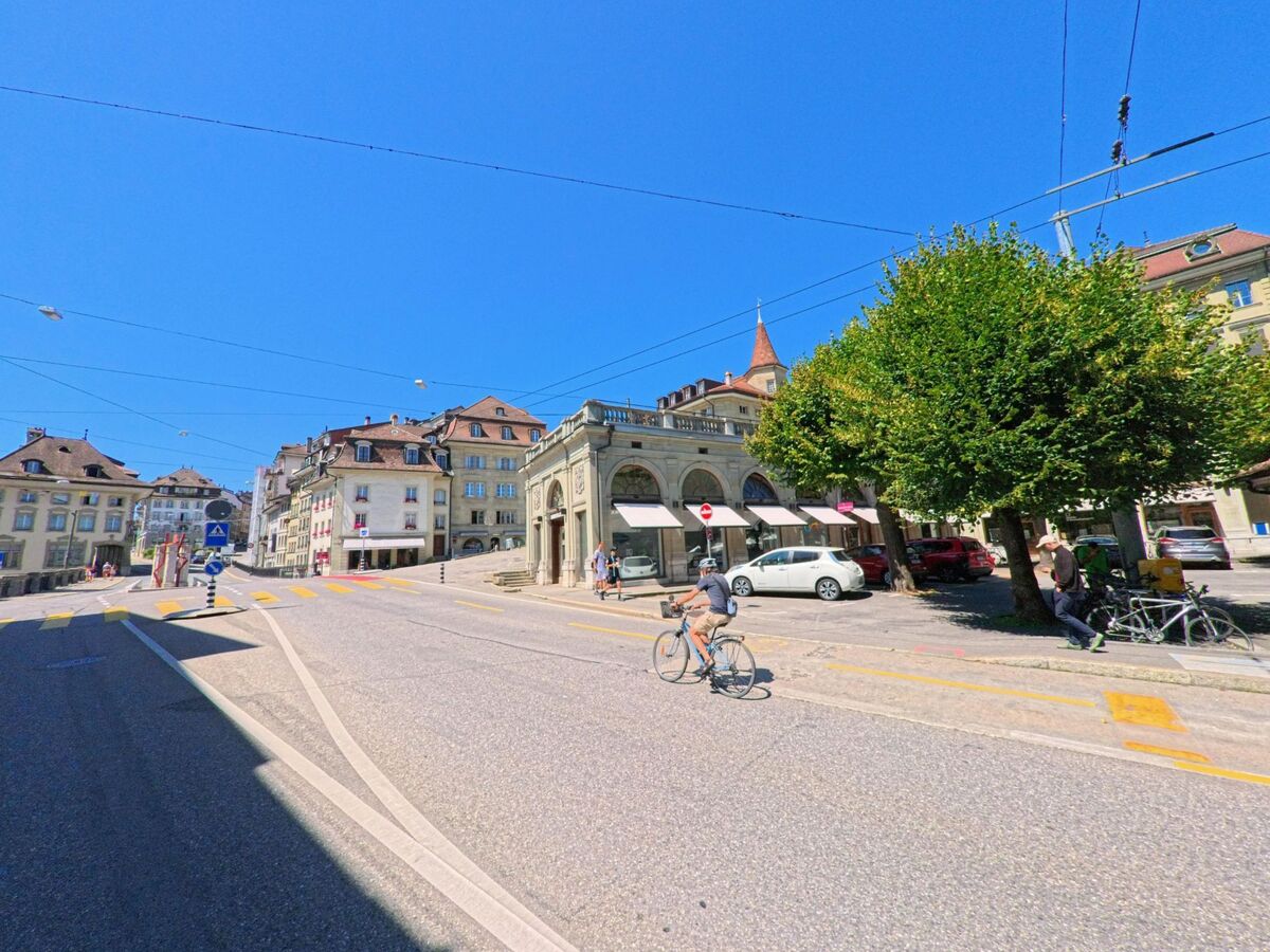 Eine belebte Strasse in Fribourg (1700, Kanton Fribourg) zeigt Radfahrer und Fussgaenger vor historischen Haeusern mit Arkaden und einem markanten Turm.