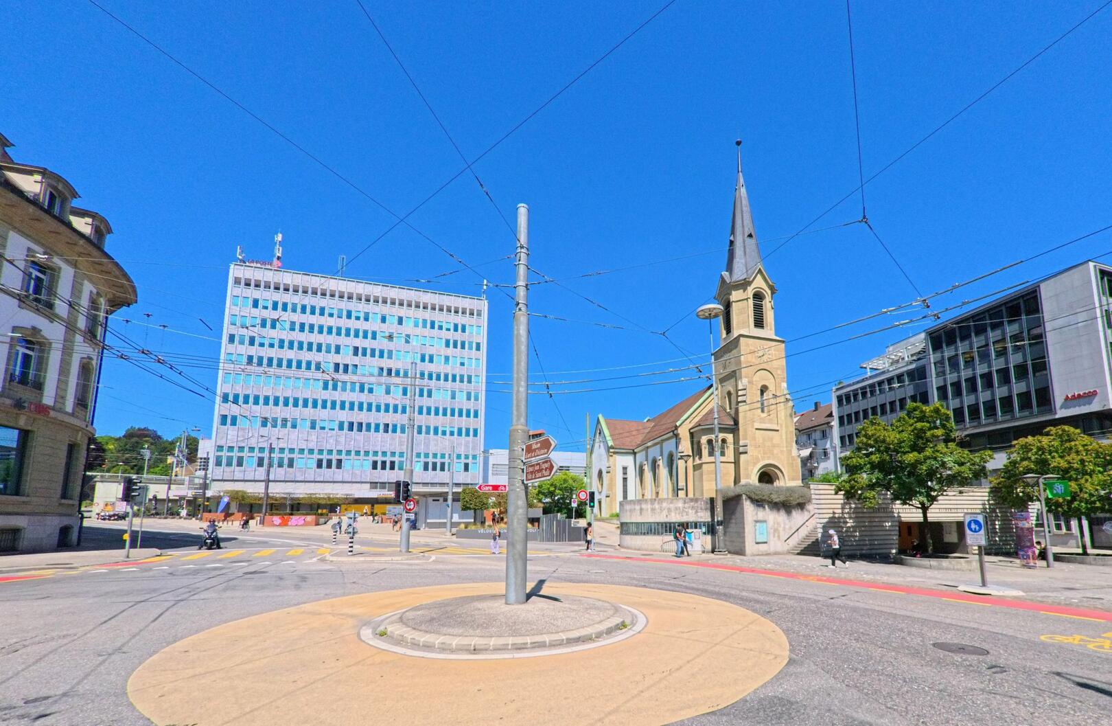 An einer Verkehrsinsel in Fribourg (1700, Kanton Fribourg) steht eine Kirche mit hohem Turm neben modernen Verwaltungsgebaeuden.