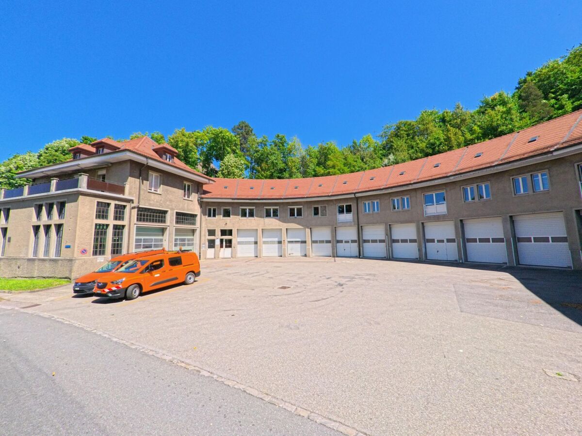 Ein halbkreisfoermiges Gebaeude mit zahlreichen Garagentoren und roten Daechern steht in Fribourg (1722, Kanton Freiburg) neben einer Waldkante.