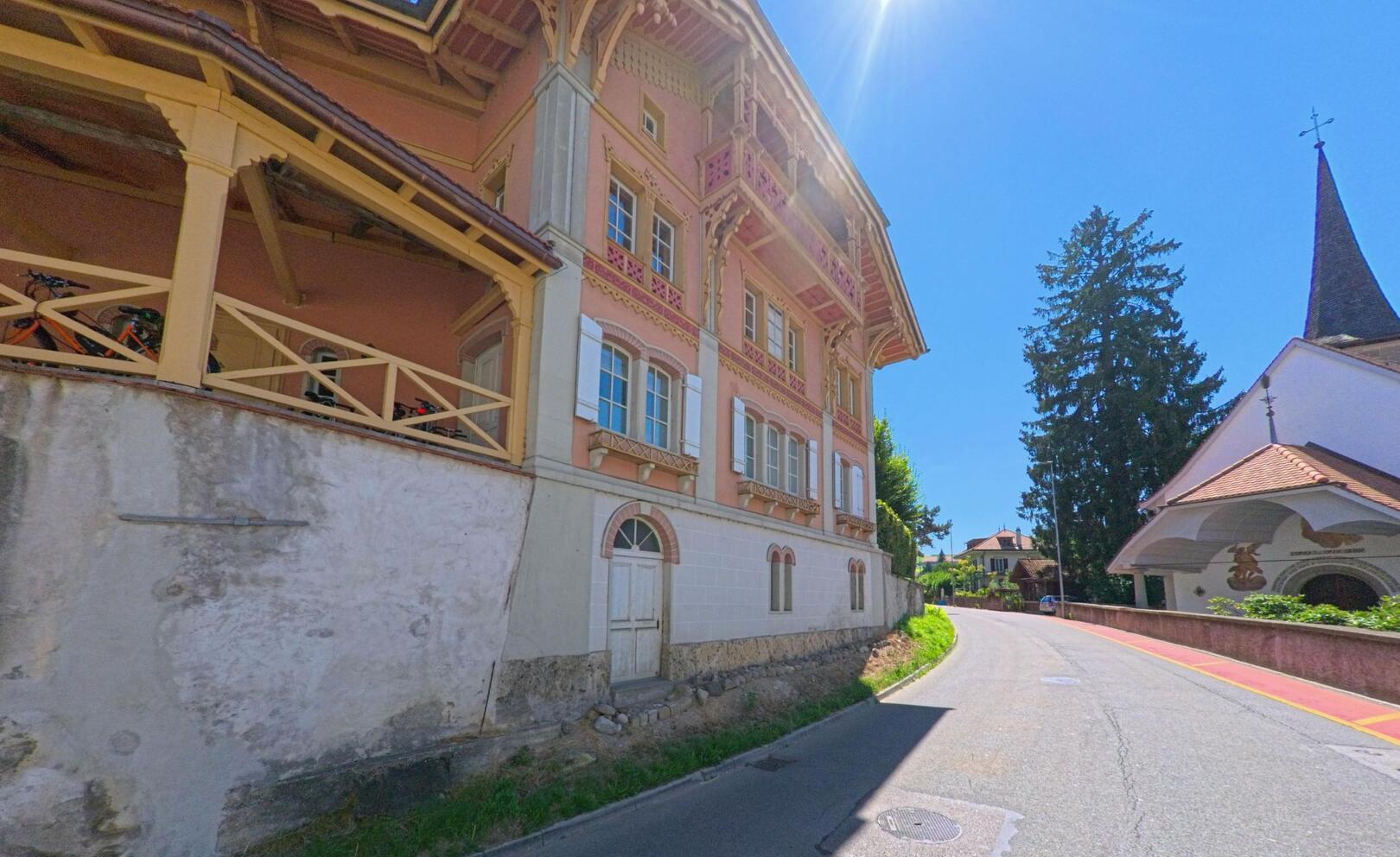 Eine schmale Strasse in Fribourg (1722, Kanton Freiburg) fuehrt zwischen einer Kirche mit spitzem Turm und einem verzierten Wohnhaus bergauf.