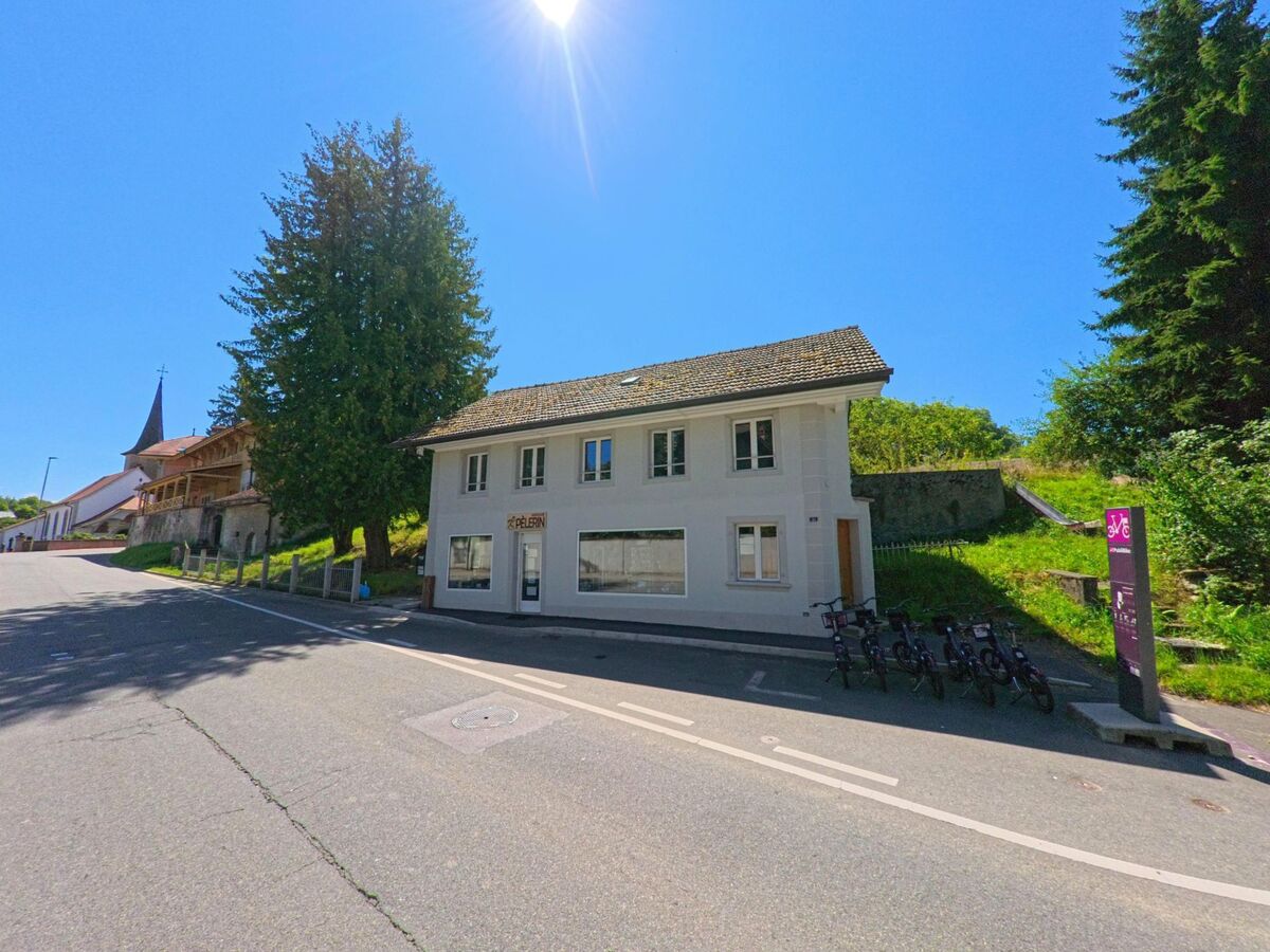Ein schlichtes weisses Haus mit Schaufenstern steht in Fribourg (1722, Kanton Freiburg) an einer Strasse mit Veloverleihstation, im Hintergrund eine Kirche.
