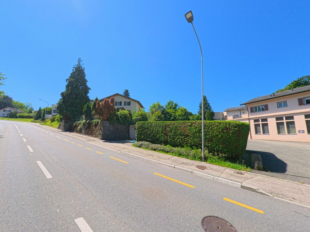 Eine leicht ansteigende Strasse in Fribourg (1722, Kanton Freiburg) fuehrt vorbei an Wohnhaeusern mit Gaerten und einem Gebaeude mit rosa Fassade.