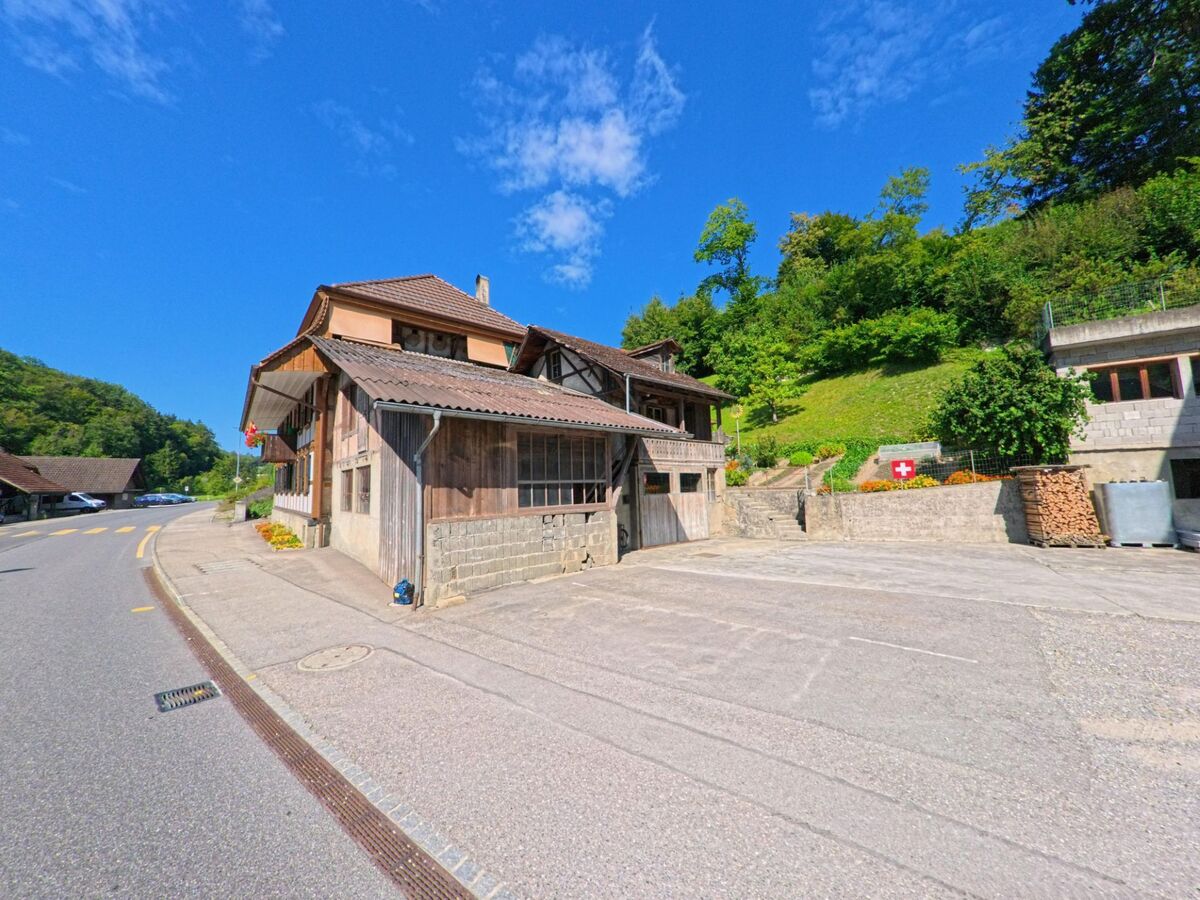 Ein traditionelles Bauernhaus mit Holzfassade steht in Fribourg (3176, Kanton Freiburg) an einer leicht kurvigen Strasse nahe einem bewaldeten Hang.