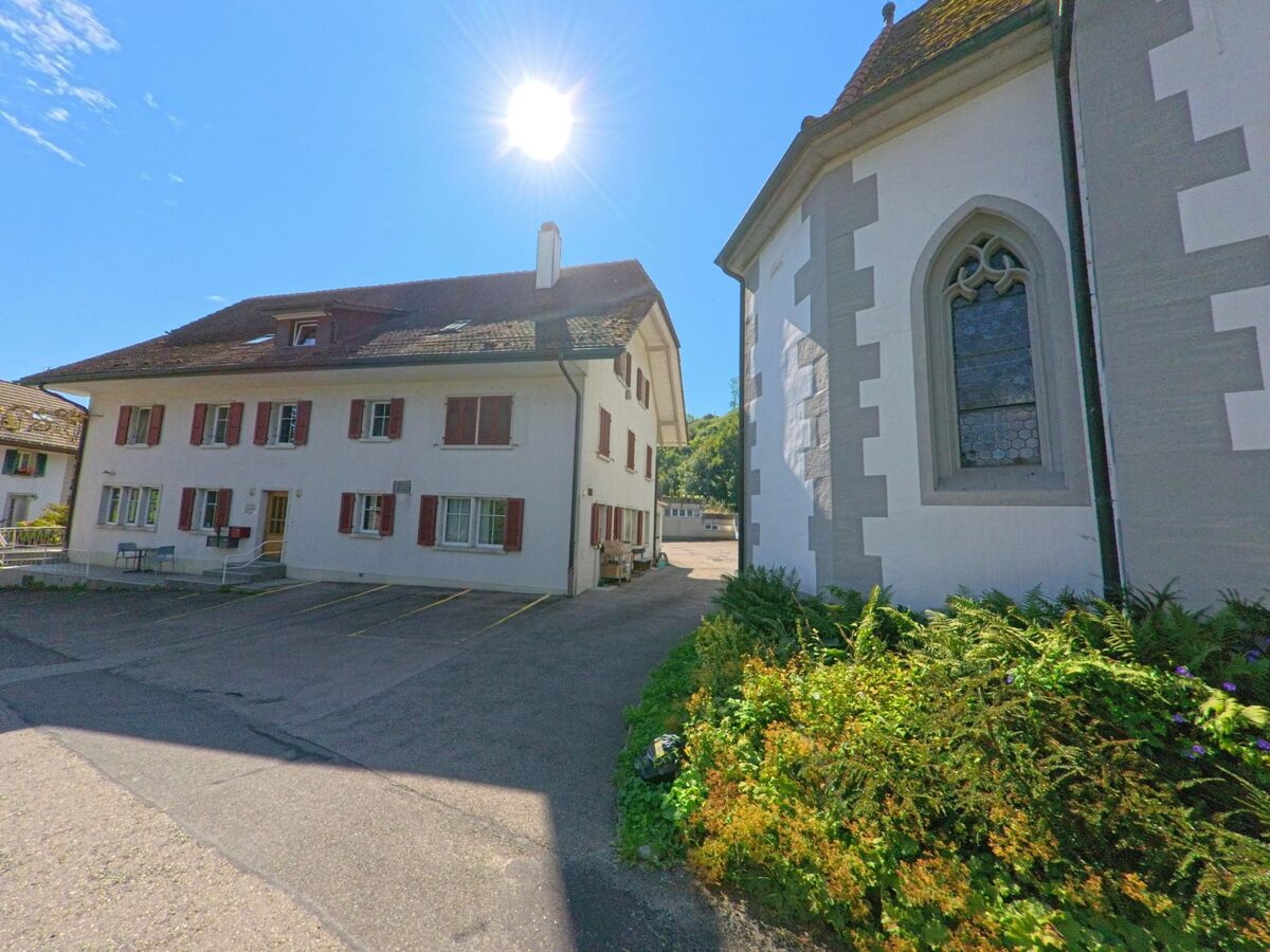 Neben einer Kirche mit Spitzbogenfenster steht in Fribourg (3176, Kanton Freiburg) ein weisses Wohnhaus mit roten Fensterlaeden und kleinem Vorplatz.