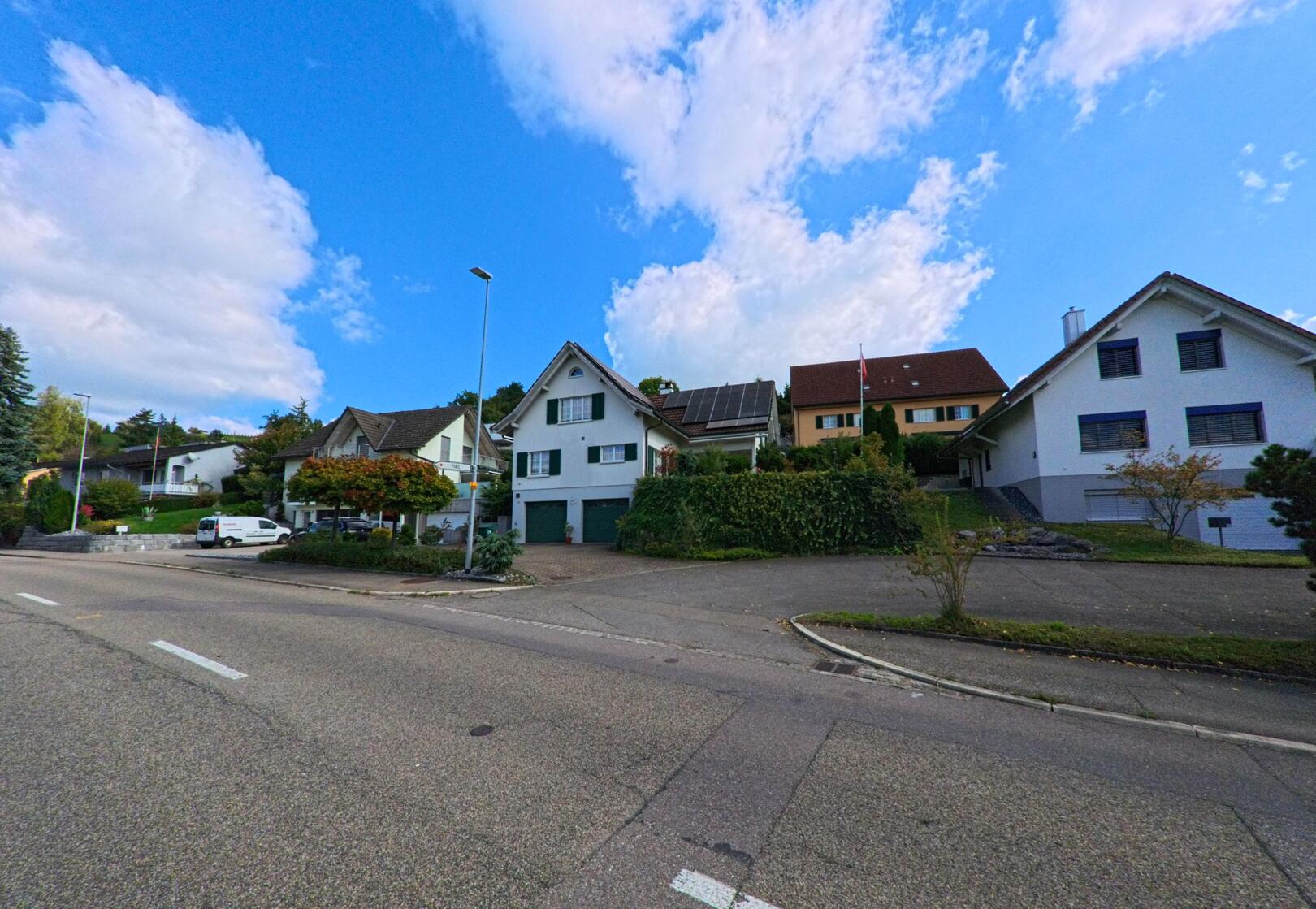 Mehrere Einfamilienhaeuser mit gepflegten Gaerten liegen an einer leicht ansteigenden Strasse in Frick (5070, Kanton Aargau) unter einem blauen Himmel mit lockeren Wolken.