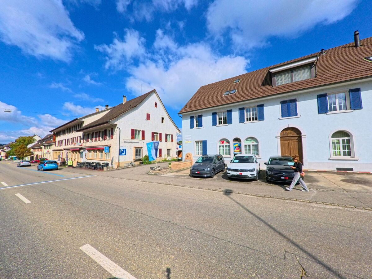 An der Hauptstrasse in Frick (5070, Kanton Aargau) stehen traditionelle Gebaeude mit farbigen Fensterlaeden, vor denen Autos parken und ein Mensch die Strasse ueberquert.
