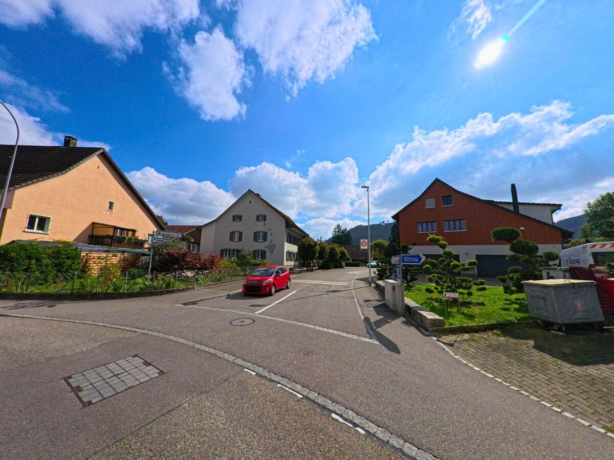 Eine ruhige Strassenkreuzung in Frick (5070, Kanton Aargau) zeigt Wohnhaeuser mit gepflegten Gaerten und einen Wegweiser in Richtung Wittnau unter strahlend blauem Himmel.