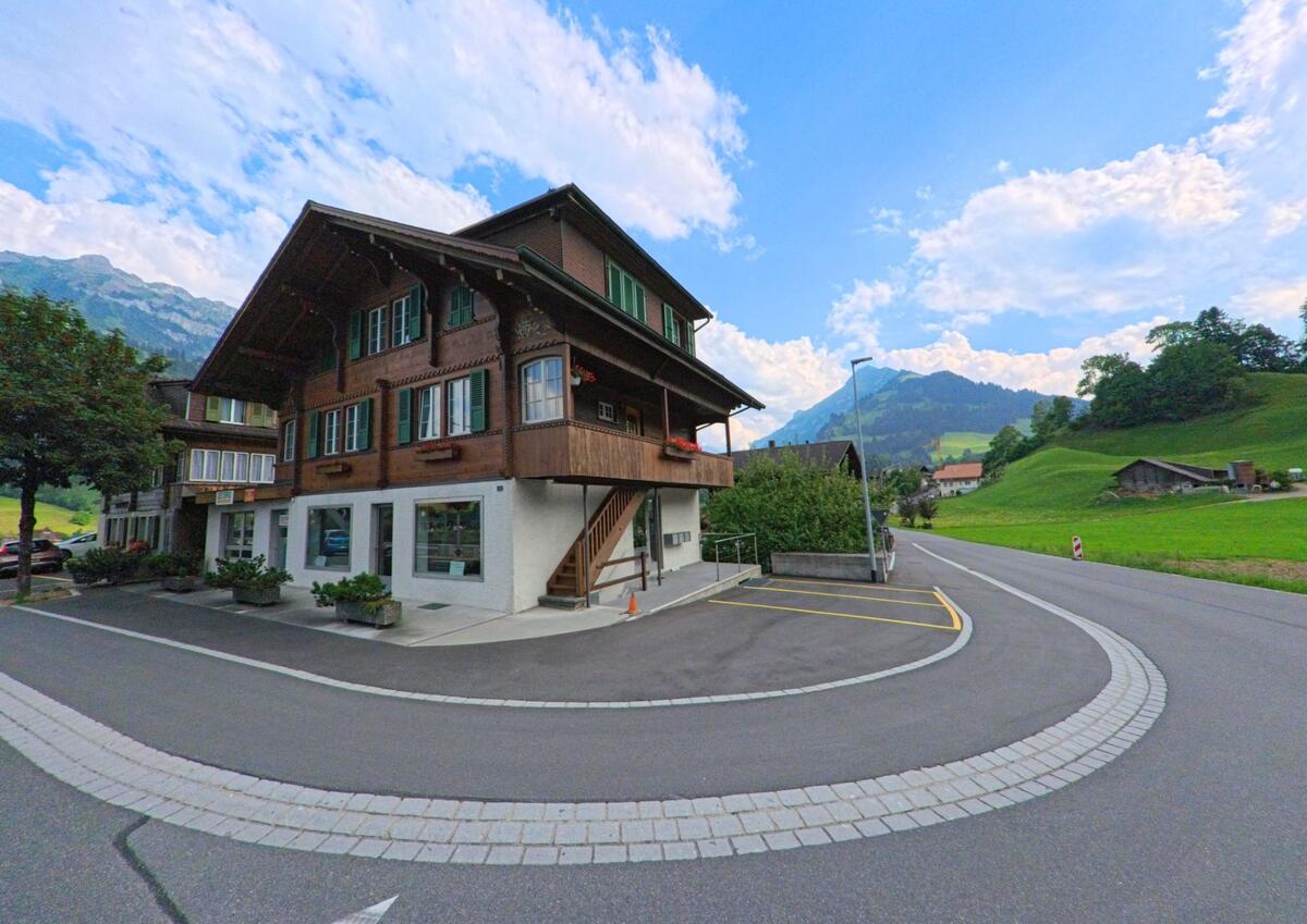 Am Übergang zur offenen Landschaft steht in Frutigen (3714, Kanton Bern) ein traditionsreiches Holzhaus mit grünen Fensterläden und Blick auf die umliegenden Hügel.