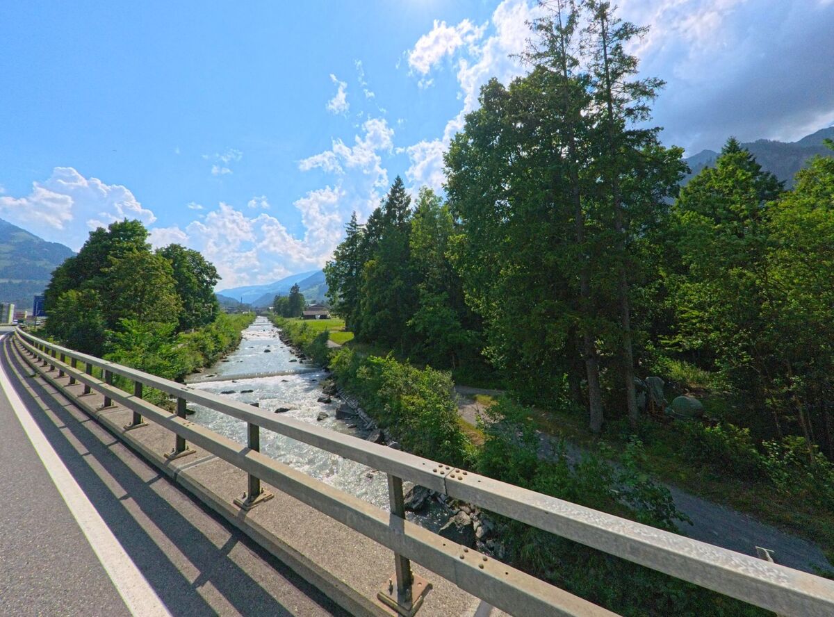 In Frutigen (3714, Kanton Bern) fliesst ein klarer Gebirgsbach neben einer Strassenbrücke durch das grüne Tal, begleitet von Bäumen und Blick auf die umliegenden Berge.