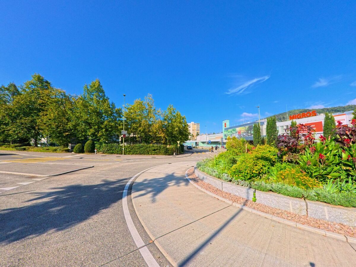 In Füllinsdorf (4414, Kanton Basel-Landschaft) liegt ein Einkaufsareal mit Migros-Gebäude hinter einem bunt bepflanzten Kreisel, während eine Baumreihe die Strasse säumt.