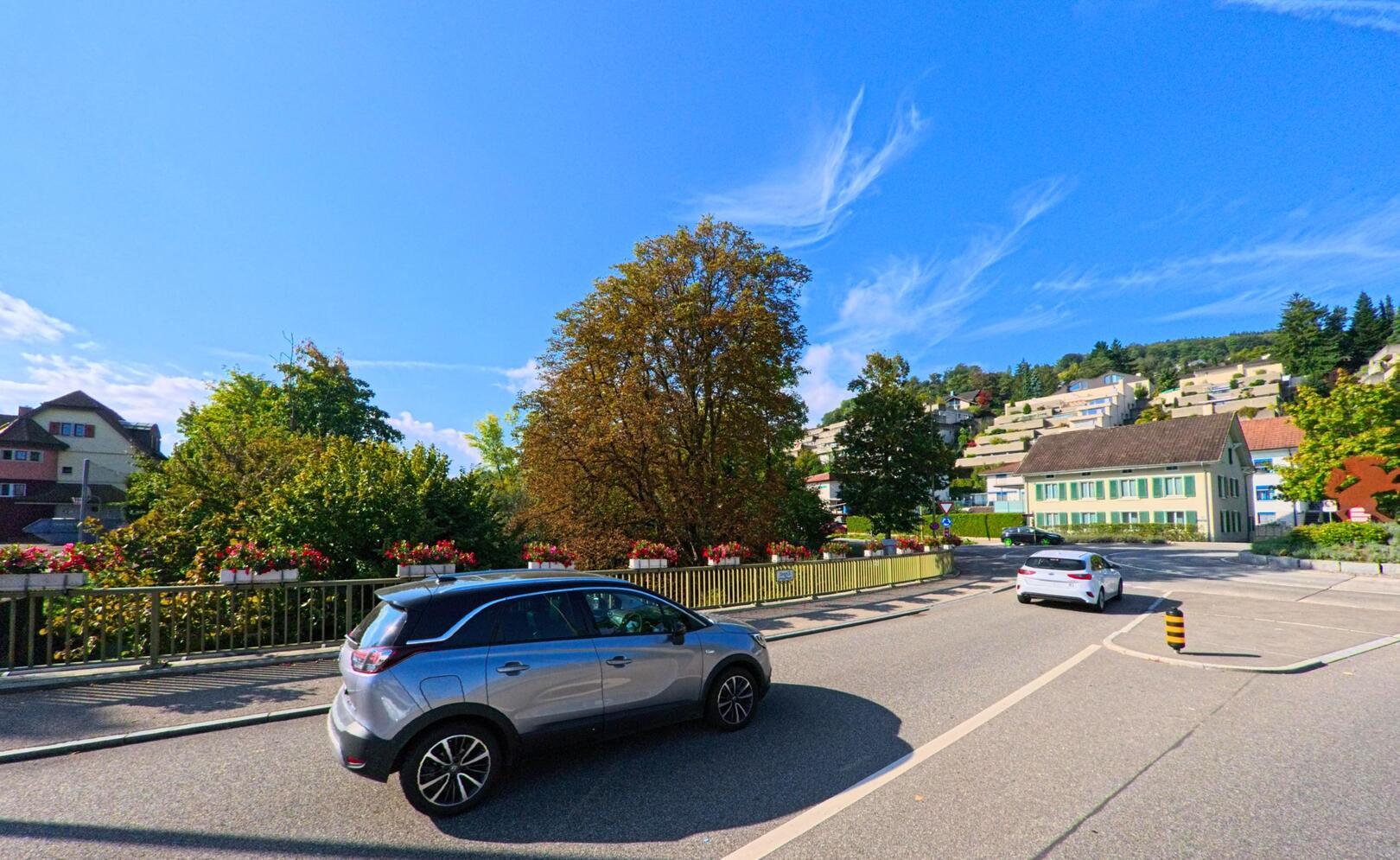 Auf einer Kreuzung in Füllinsdorf (4414, Kanton Basel-Landschaft) fahren zwei Autos an bepflanzten Brückengeländern vorbei, während über den Hang hinweg Wohnbauten in der Sonne liegen.