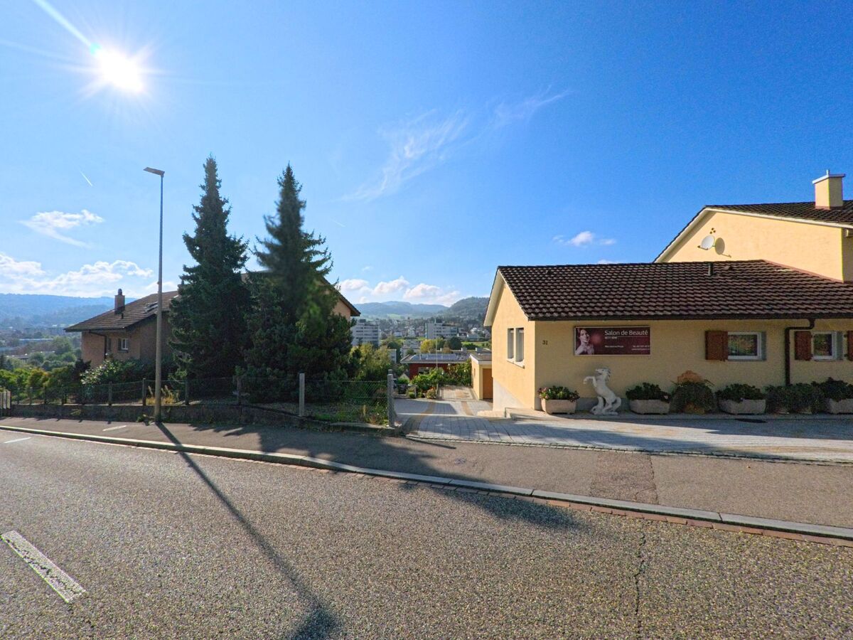 Vor einem gelben Wohnhaus mit einem kleinen Schönheitssalon in Füllinsdorf (4414, Kanton Basel-Landschaft) öffnet sich der Blick über die Siedlung bis zu den entfernten Hügeln.
