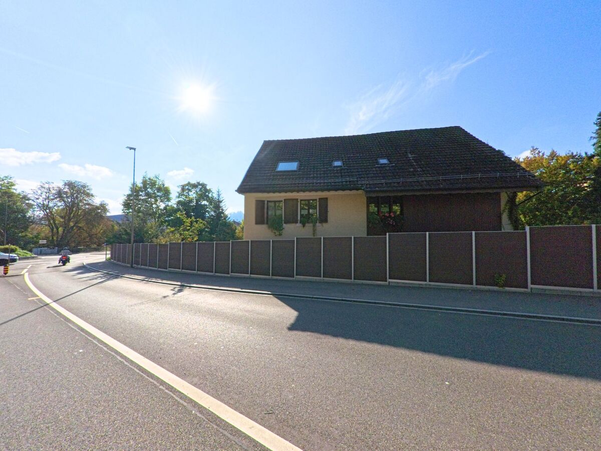 An einer kurvigen Strasse in Füllinsdorf (4414, Kanton Basel-Landschaft) steht ein Wohnhaus hinter einer hohen Schallschutzwand, während ein Motorradfahrer in der Sonne vorbeifährt.