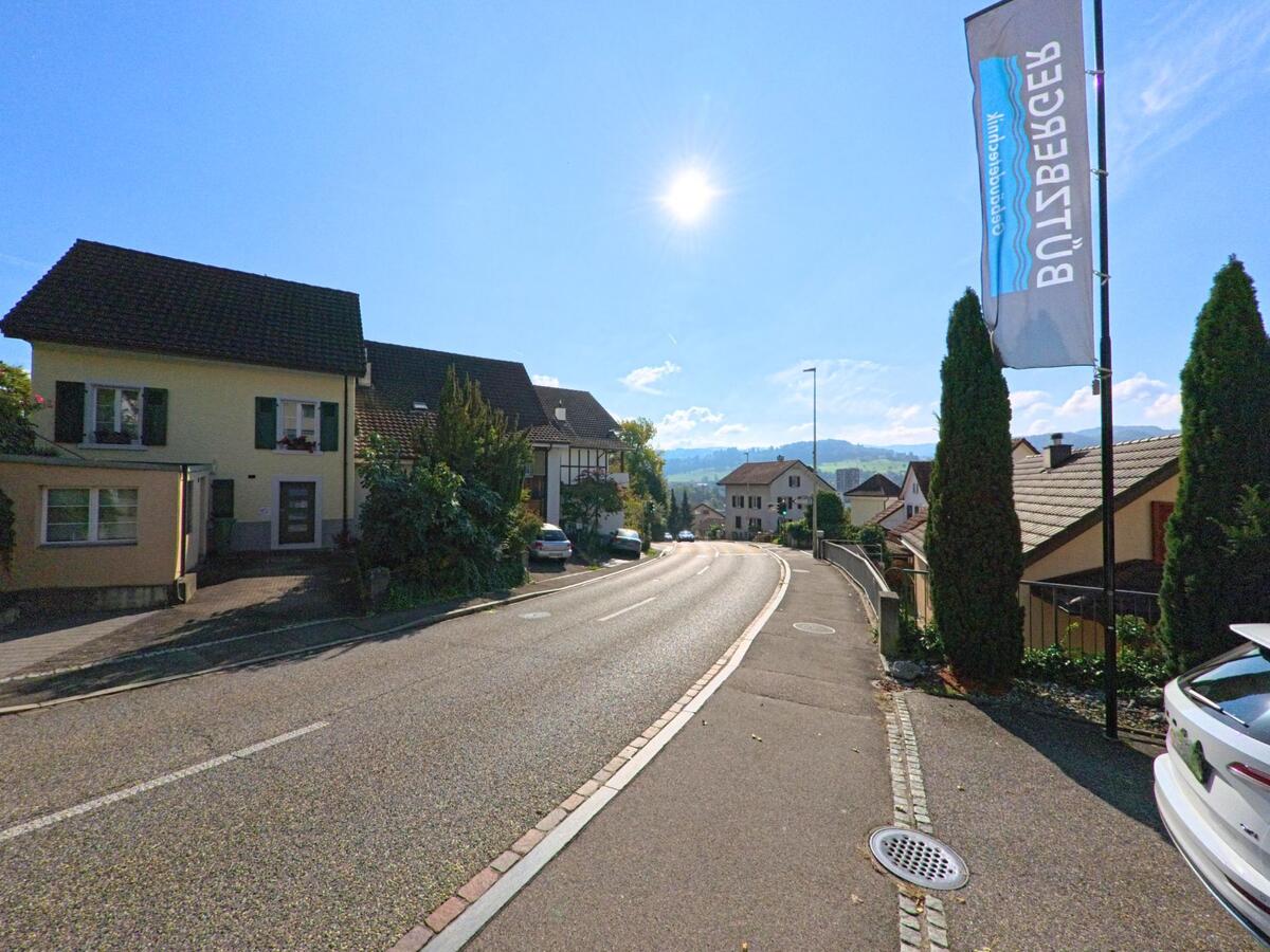 Eine leicht ansteigende Wohnstrasse in Füllinsdorf (4414, Kanton Basel-Landschaft) zeigt Häuser mit Vorgärten und weiten Blick auf die hügelige Landschaft unter der tief stehenden Sonne.