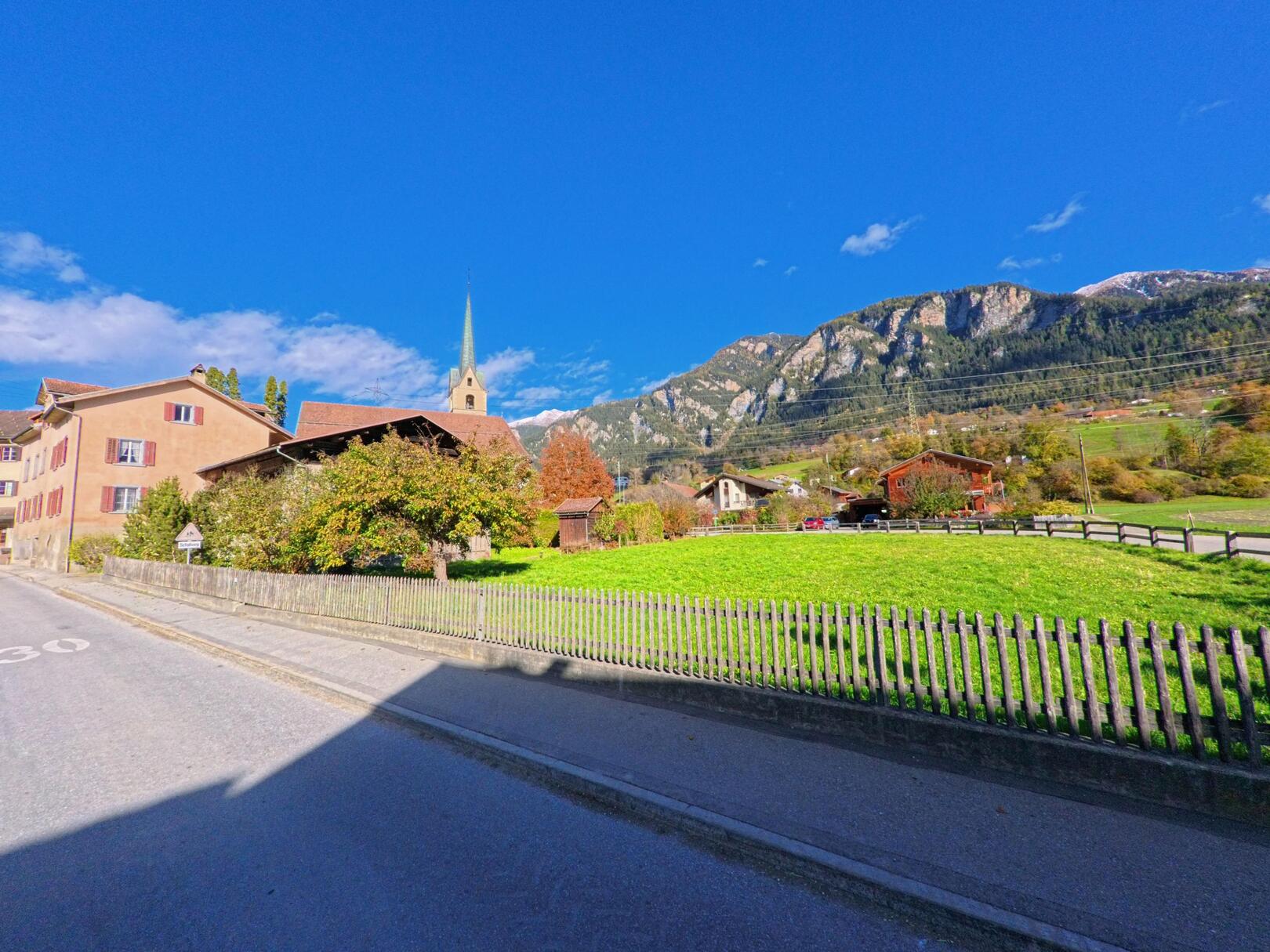 Entlang einer ruhigen Strasse öffnet sich der Blick über eine grüne Wiese mit Holzzaun auf Wohnhäuser und den Kirchturm von Fürstenau (7414, Kanton Graubünden), vor der Kulisse felsiger Berghänge.