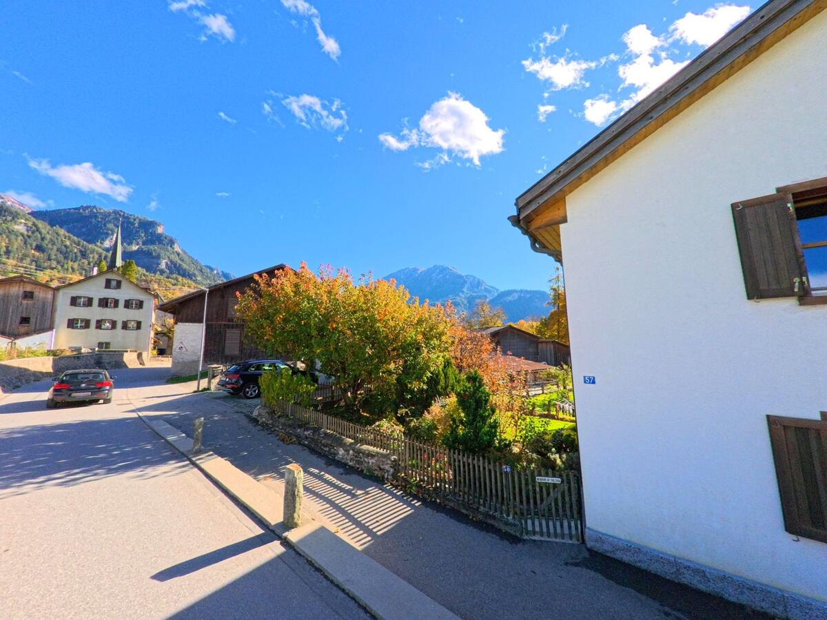 Zwischen Wohnhäusern und einem gepflegten Garten verläuft eine ruhige Strasse in Fürstenau (7414, Kanton Graubünden), begleitet von herbstlicher Vegetation und Bergen im Hintergrund.