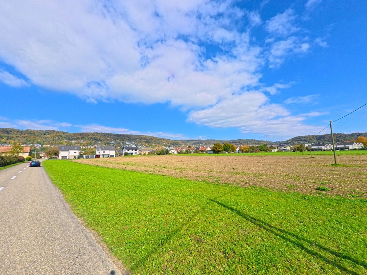 Ein Auto faehrt auf einer Landstrasse durch Full-Reuenthal (5324, Kanton Aargau) entlang weiter Felder mit Blick auf Wohnhaeuser und bewaldete Huegel.
