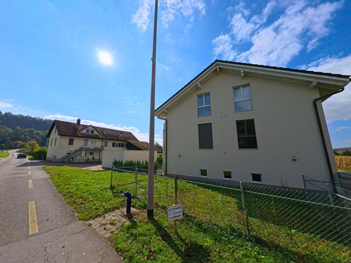 Zwei helle Wohnhaeuser in Full-Reuenthal (5324, Kanton Aargau) stehen an einer sonnigen Strasse mit Blick auf Felder und bewaldete Huegel.