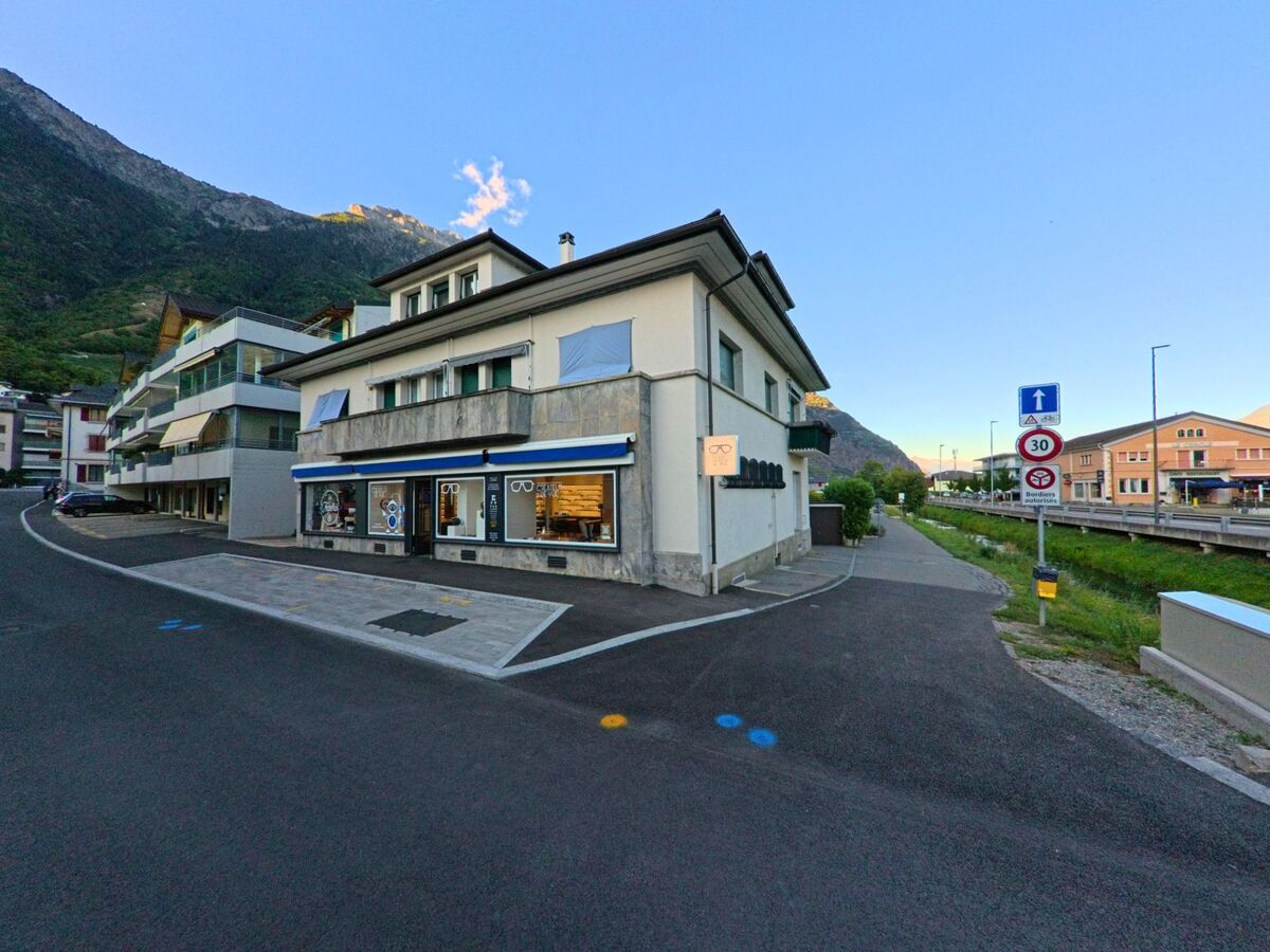 Ein Geschaeftshaus mit Schaufenstern fuer Optik befindet sich in Fully (1926, Kanton Wallis) an einer Strasse mit Verkehrsschildern und Blick auf die Berge.