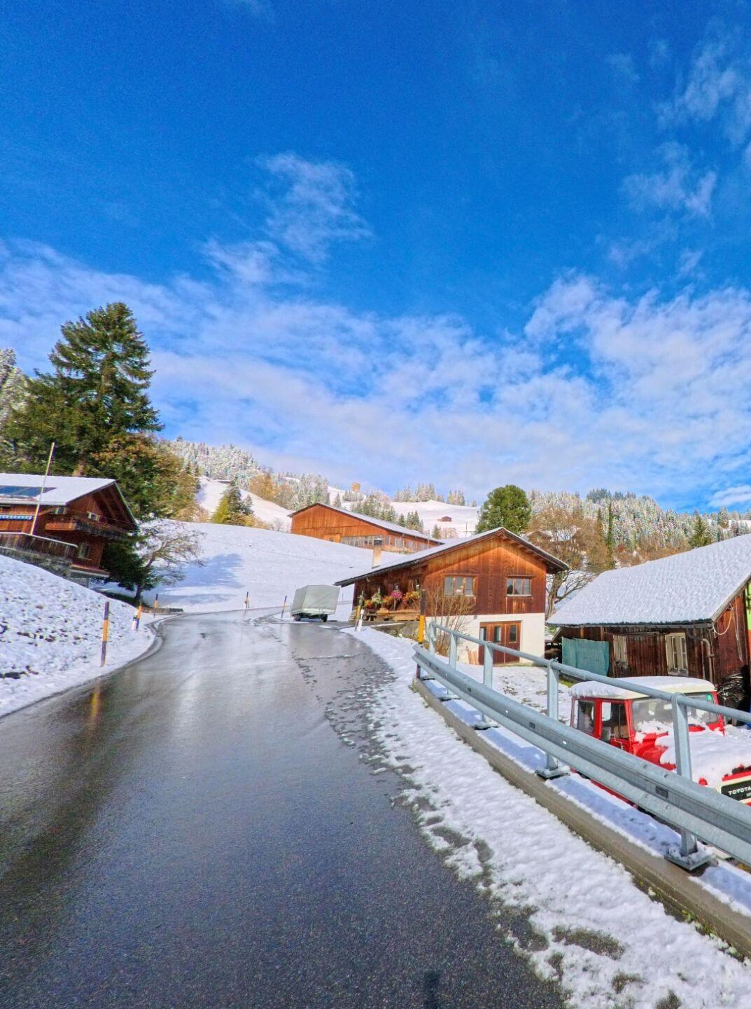 Entlang einer ansteigenden Dorfstrasse mit Schneeresten führt der Blick auf mehrere Holzhäuser in Furna (7232, Kanton Graubünden), eingerahmt von bewaldeten Hängen und klarem Himmel.
