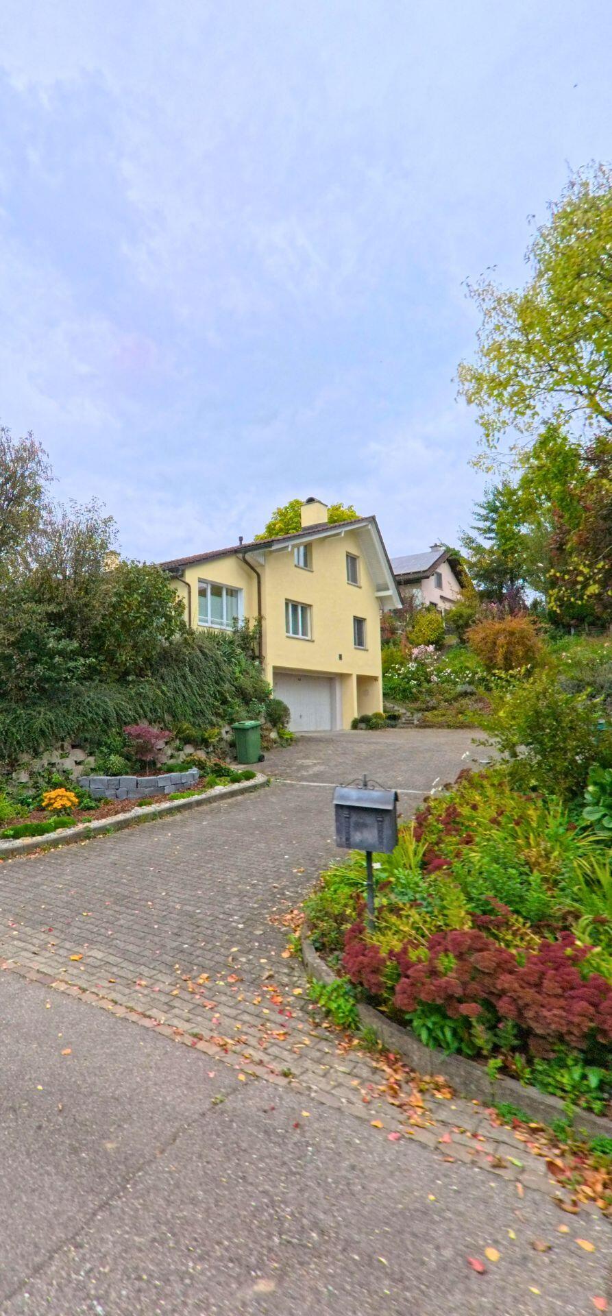 Ein Einfamilienhaus mit Einfahrt und Garage liegt zwischen üppigen Gärten in Gachnang (8547, Kanton Thurgau), während ein Briefkasten am Rand des Vorplatzes steht.