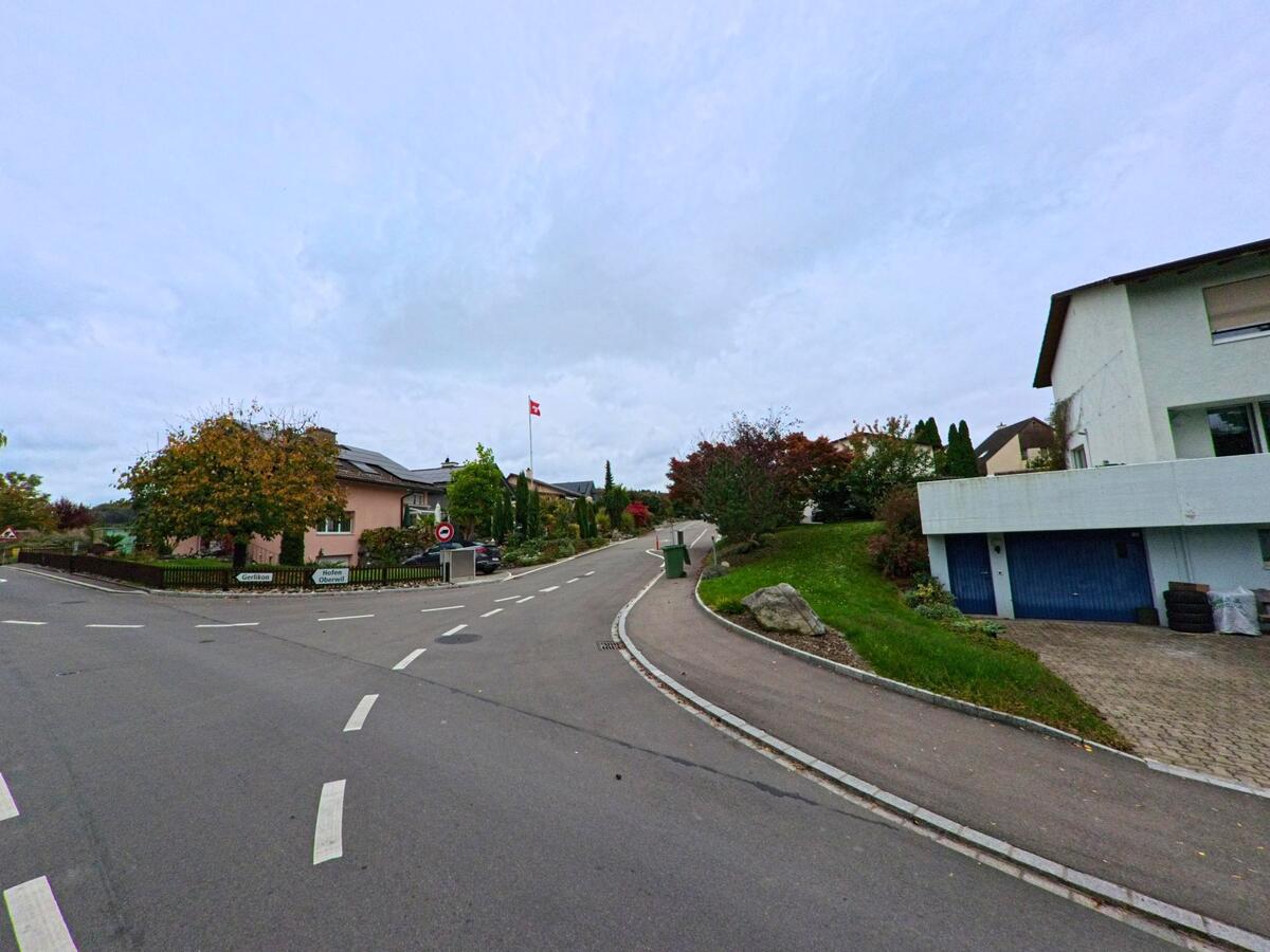 Eine sanft ansteigende Quartierstrasse mit Gehweg und Kurve führt durch ein Wohngebiet in Gachnang (8547, Kanton Thurgau), wo eine Schweizer Fahne über gepflegten Gärten weht.