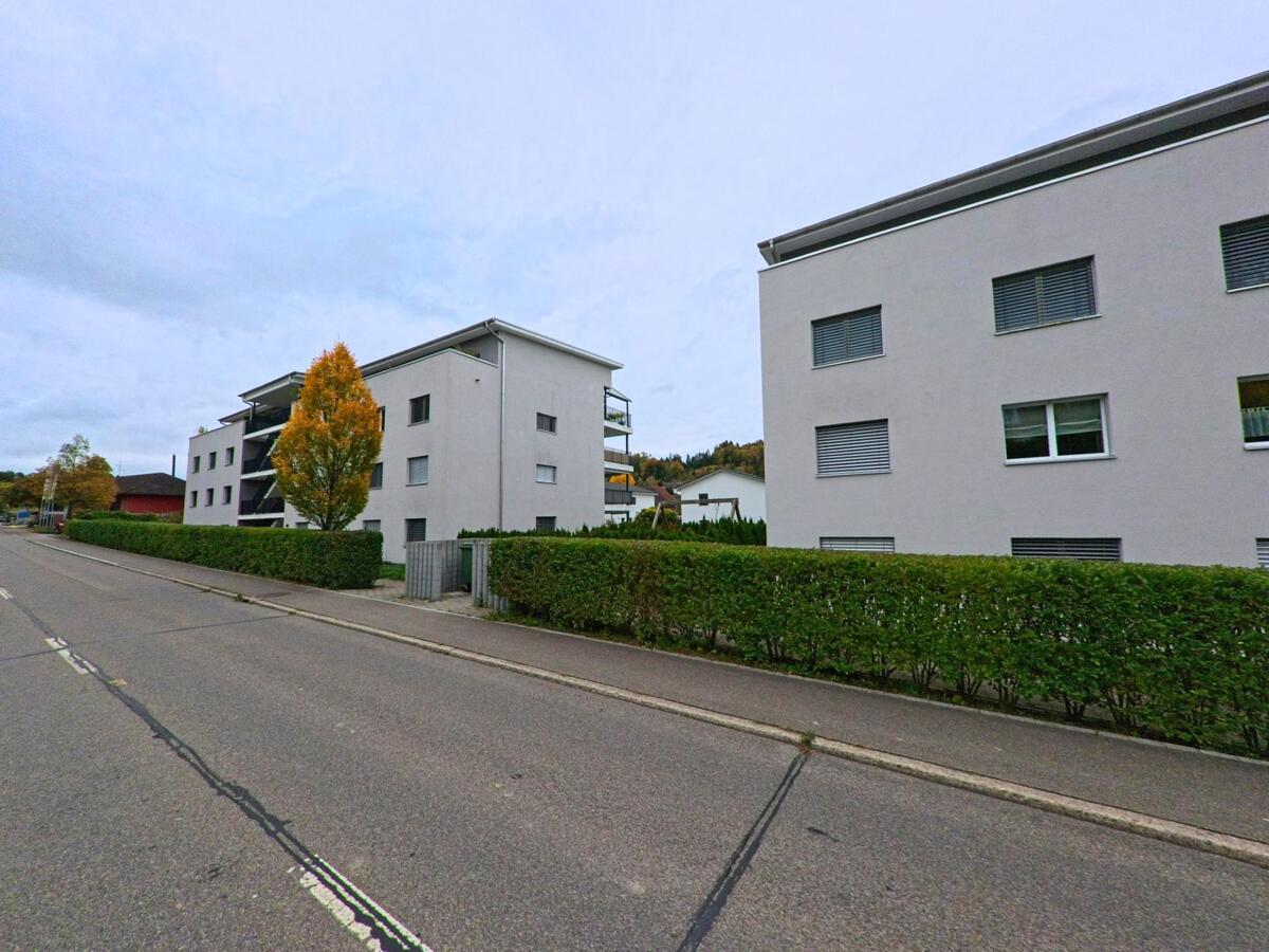 Hinter einer langen Hecke stehen helle Mehrfamilienhäuser mit Balkonen in Gachnang (8547, Kanton Thurgau), und ein Baum mit Herbstfärbung lockert die Strassenfront auf.