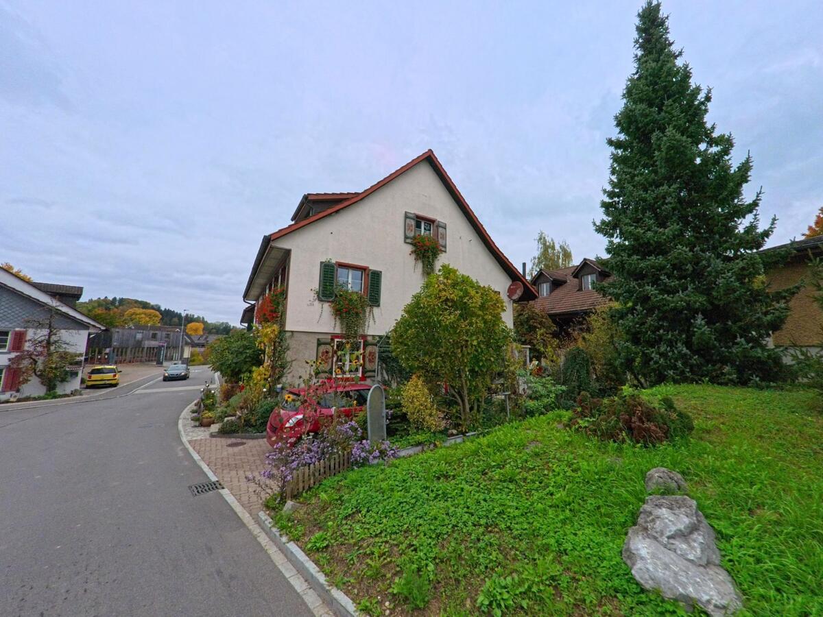 Ein traditionelles Wohnhaus mit Fensterläden und blühenden Pflanzen steht an einer Kurve in Gachnang (8547, Kanton Thurgau), daneben ragt ein hoher Nadelbaum über eine Grünfläche.