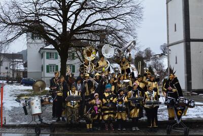 Auf einem Platz in Gais im Kanton Appenzell Ausserrhoden posiert eine verkleidete Musikgruppe mit Instrumenten vor winterlicher Kulisse und kahlem Baum.