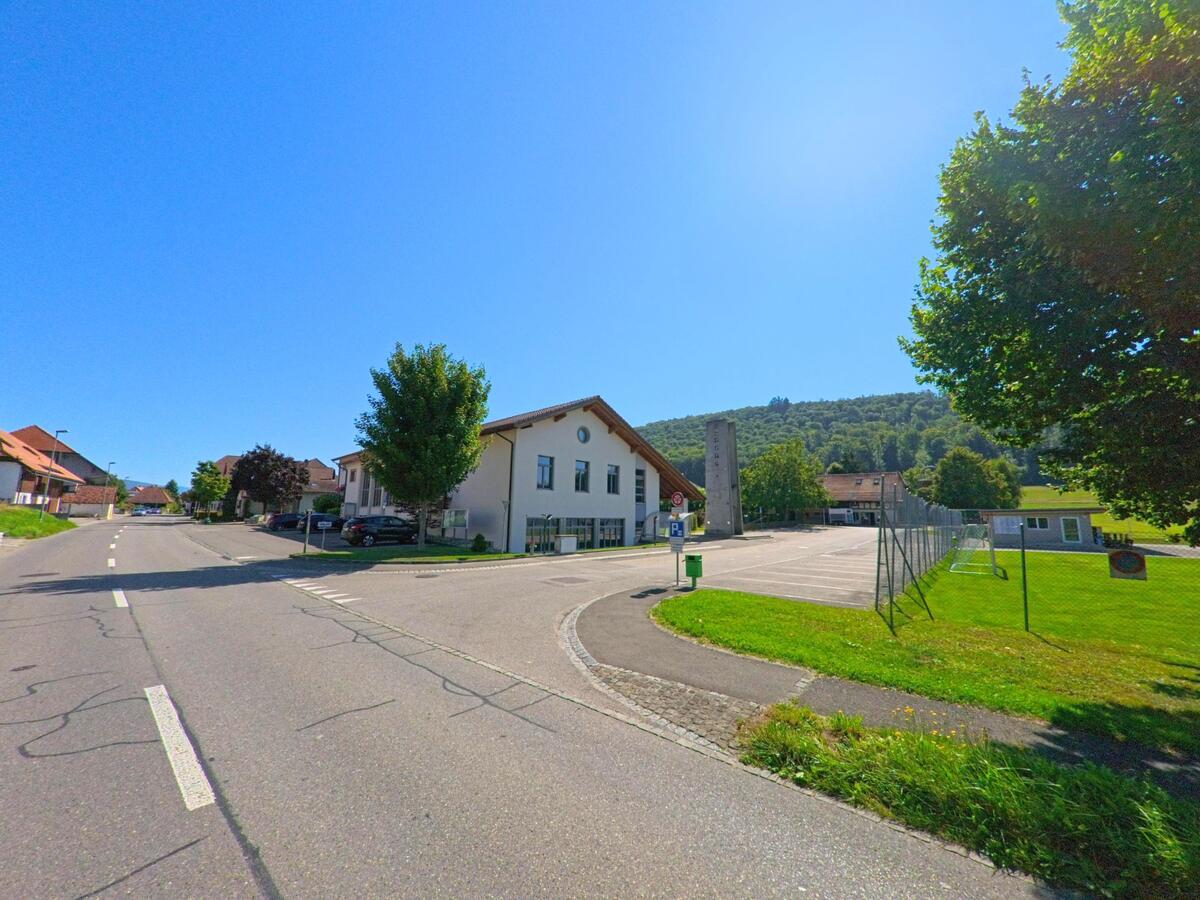 An einer Strasse in Gals (3238, Kanton Bern) steht das Schulgebaeude mit angrenzendem Sportplatz vor einem bewaldeten Hang.