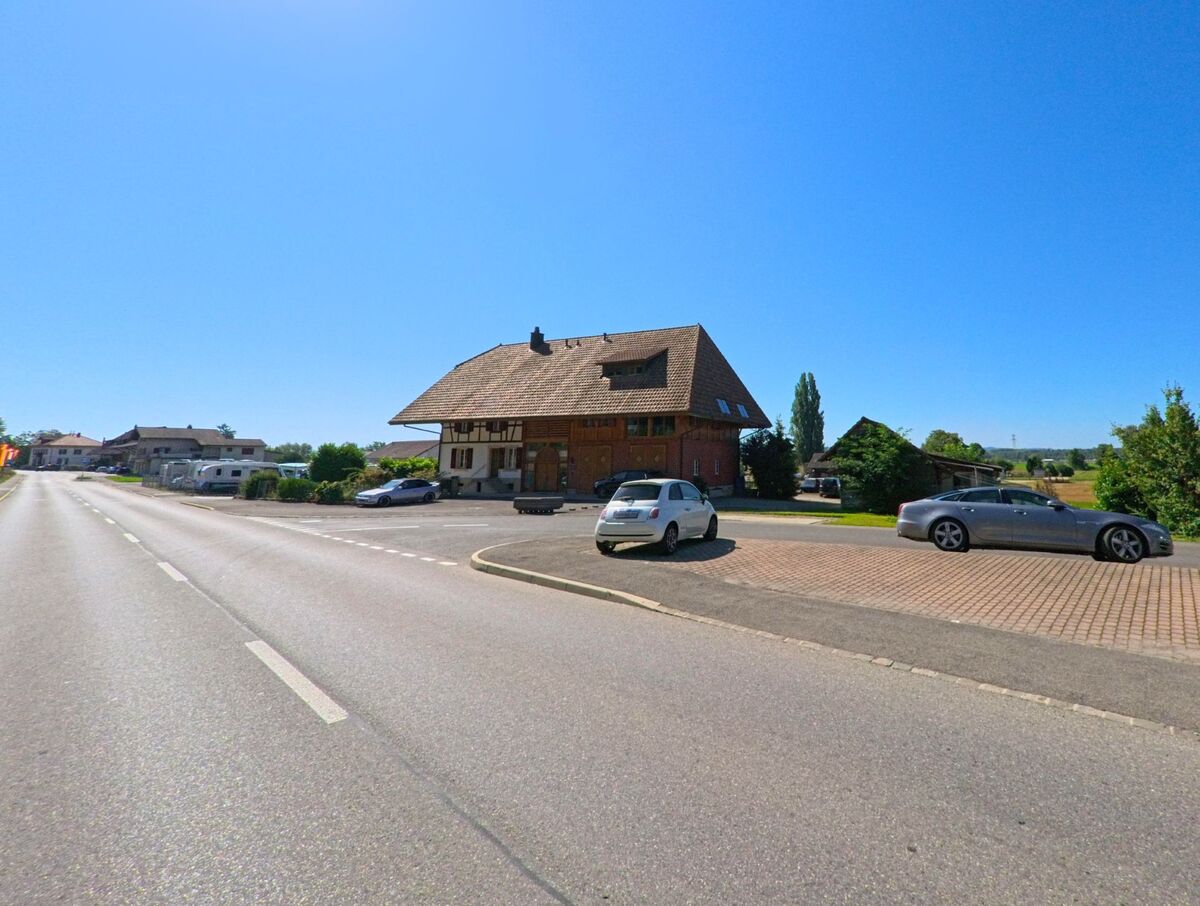 An der Hauptstrasse in Gampelen (3236, Kanton Bern) steht ein grosses Bauernhaus mit angrenzendem Parkplatz und Blick auf die umliegenden Felder.
