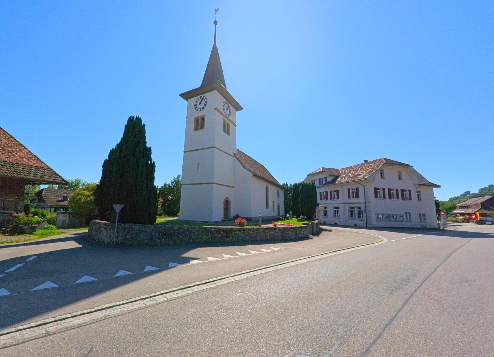 Bei strahlendem Sonnenschein steht die reformierte Kirche im Zentrum von Gampelen (3236, Kanton Bern) neben dem Gemeindehaus und weiteren Gebäuden.