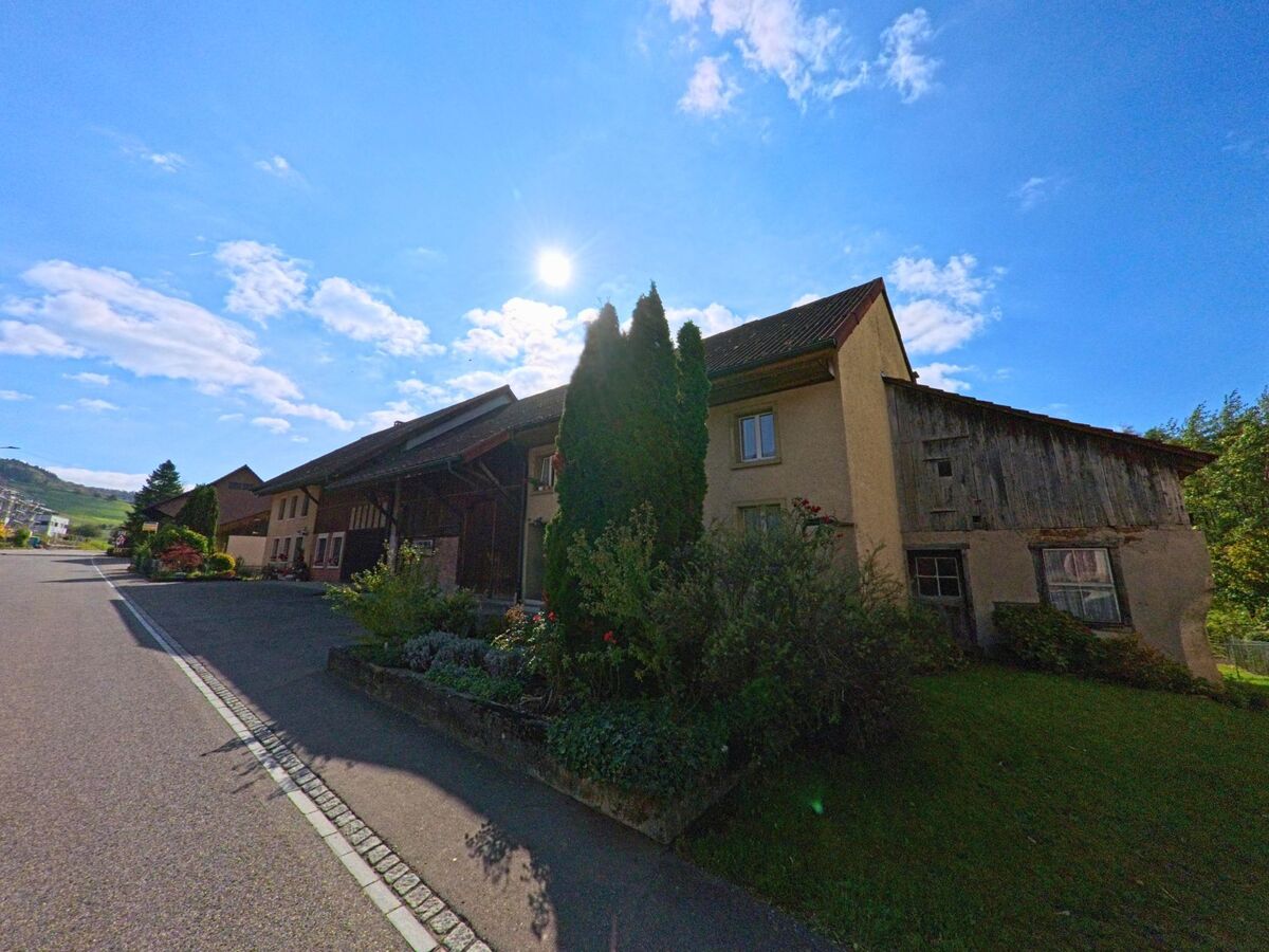 Im Zentrum von Gansingen (5272, Kanton Aargau) liegt ein kleiner Platz mit Sitzgelegenheiten, Parkplätzen und Geschäften unter blauem Himmel.