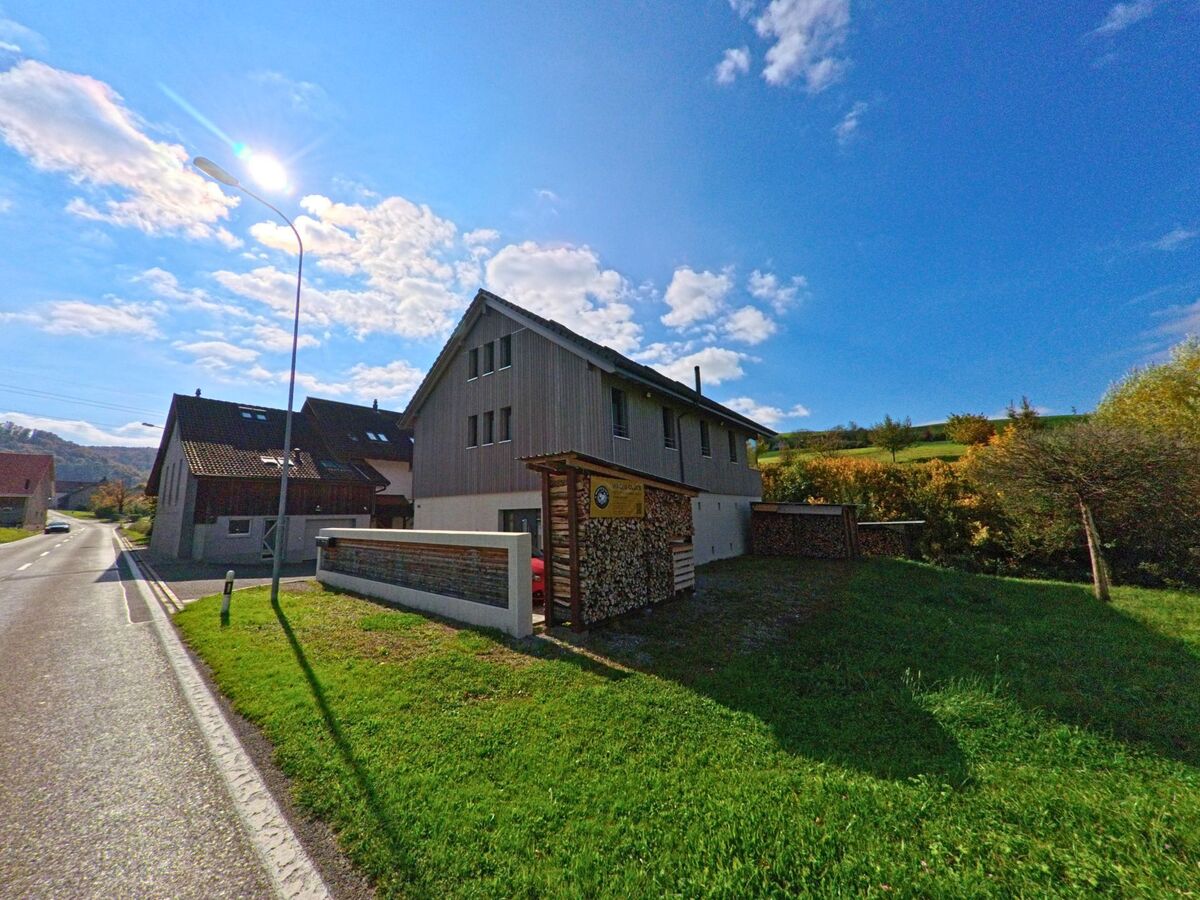 Am Dorfrand von Gansingen (5272, Kanton Aargau) steht ein modernes Einfamilienhaus mit Holzverschalung und gestapeltem Brennholz neben einer sonnigen Strasse.