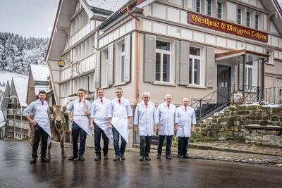 Vor dem Gasthaus und der Metzgerei Ochsen in Schwellbrunn (9103, Kanton Appenzell Ausserrhoden) posiert das Team in weissen Schuerzen mit einer Kuh.