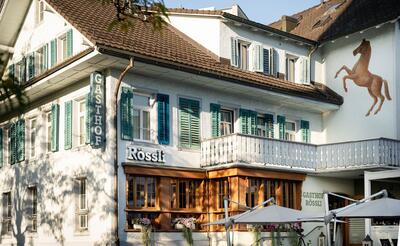 Der Gasthof Rössli in Steinhausen (6312, Kanton Zug) liegt an einer Strassenecke mit Terrasse und Sonnenschirmen, umgeben von modernen und traditionellen Gebäuden.