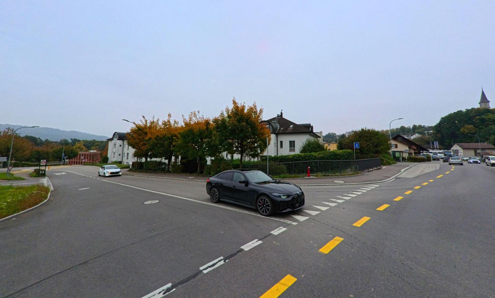 An einer Kreuzung in Gebenstorf (5412, Kanton Aargau) treffen mehrere Strassen aufeinander, im Hintergrund sind Wohnhäuser und die Kirchturmspitze der Gemeinde zu sehen.