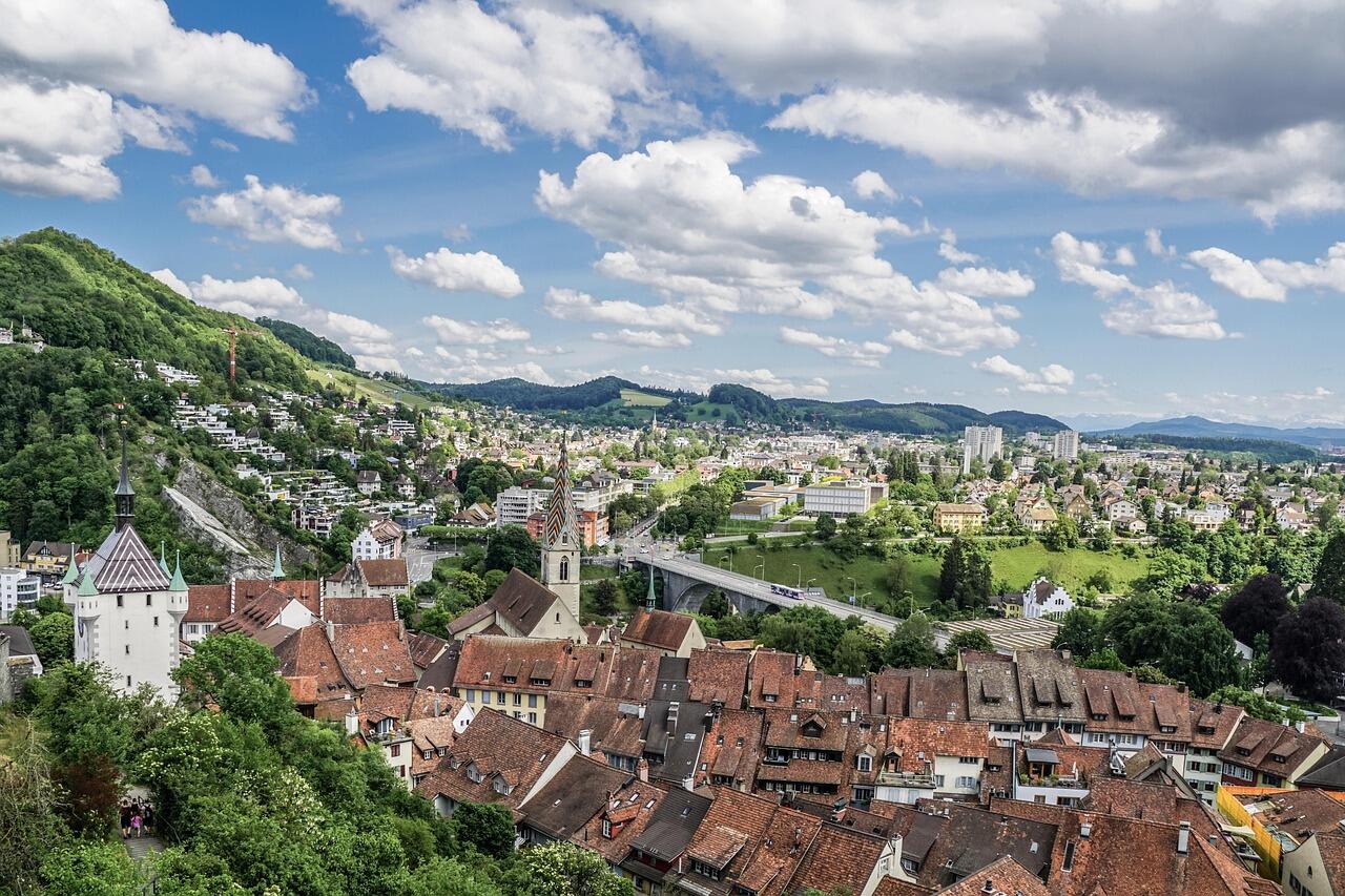 Von einer Bruecke in Baden (5415, Kanton Aargau) bietet sich ein weiter Blick auf die Haeuser am Hang und den Fluss unterhalb bei bedecktem Himmel.