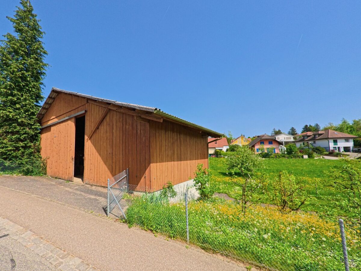 Eine einfache Holzhütte steht in Gempen (4145, Kanton Solothurn) am Rand eines Quartiers mit Einfamilienhäusern und grüner Wiese im Hintergrund.