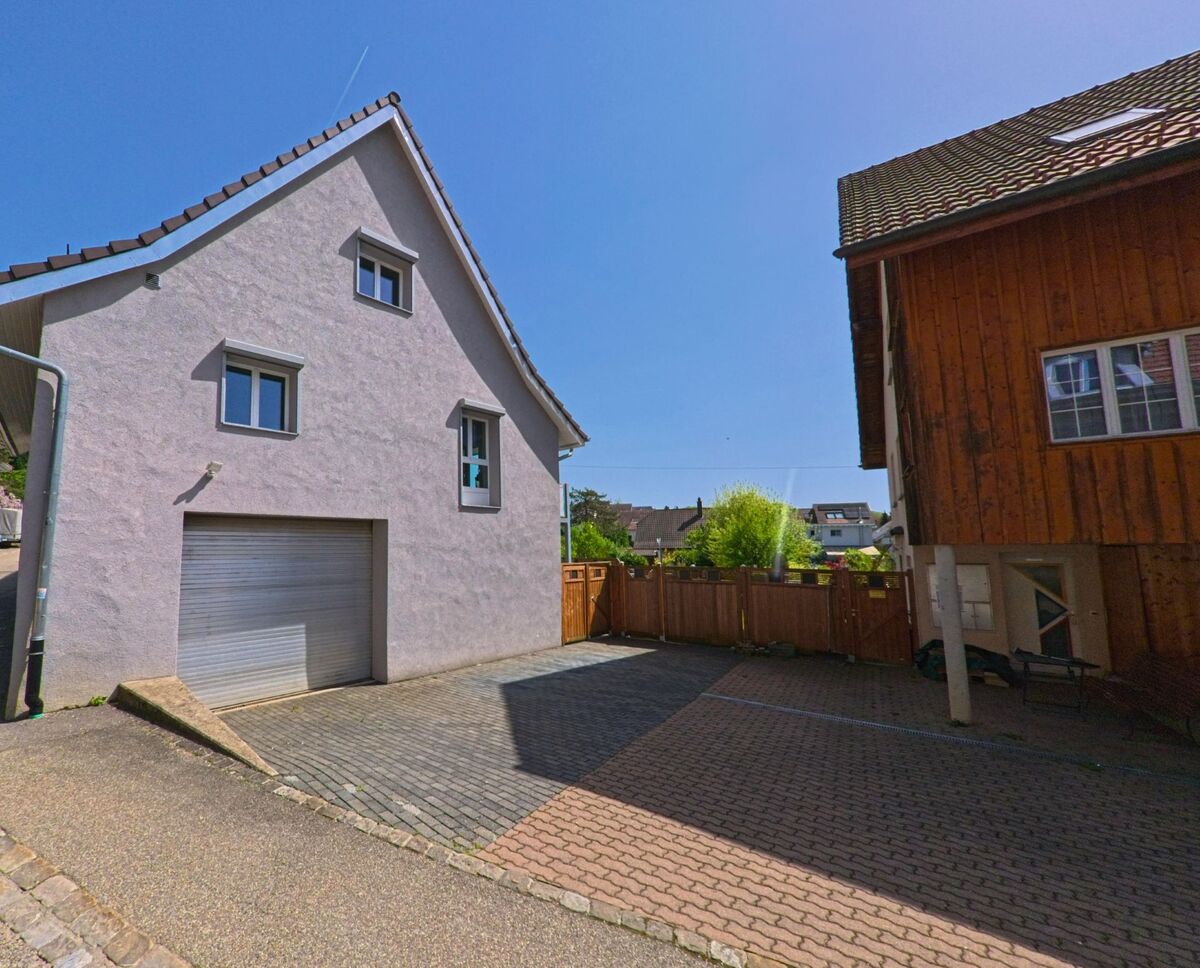 In einem Wohnquartier von Gempen (4145, Kanton Solothurn) stehen ein verputztes Haus mit Garage und ein Holzhaus gegenüber auf einem gepflasterten Vorplatz.
