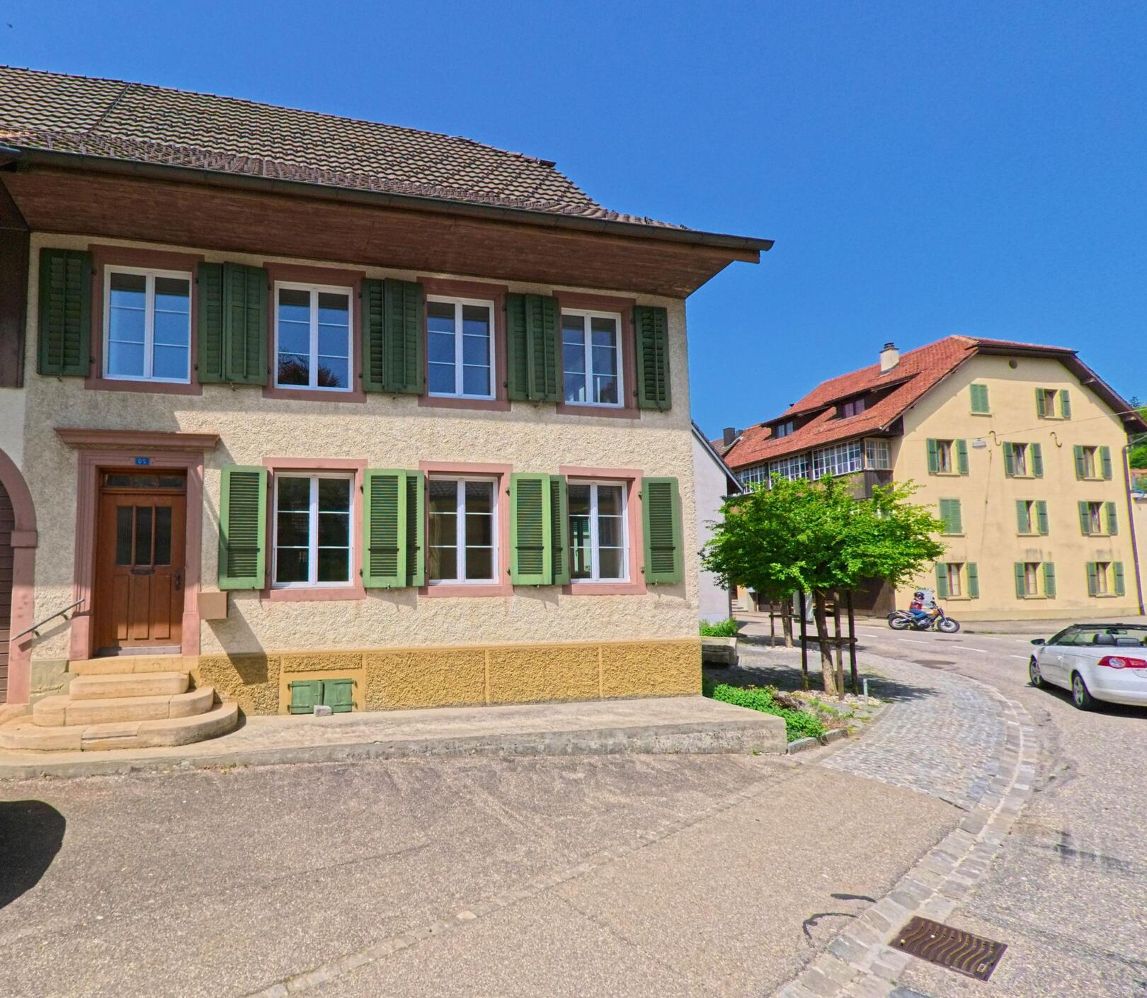 In der Gemeinde Gempen (4145, Kanton Solothurn) stehen gepflegte Wohnhäuser mit grünen Fensterläden entlang einer ruhigen Strasse bei strahlendem Sonnenschein.